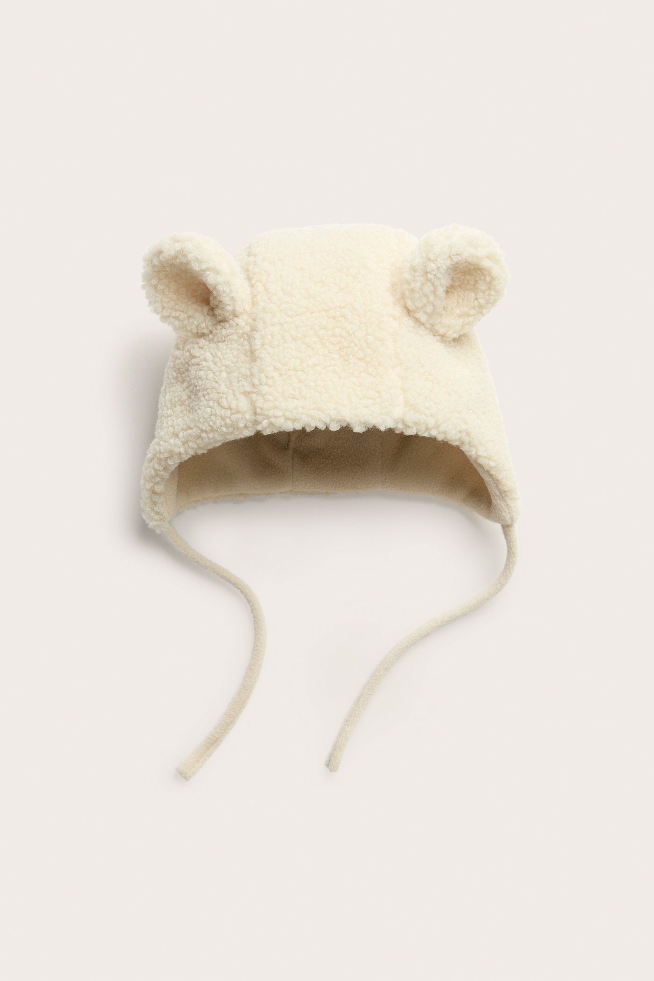 Forfra, beige babylue i pile med ører og knytebånd.