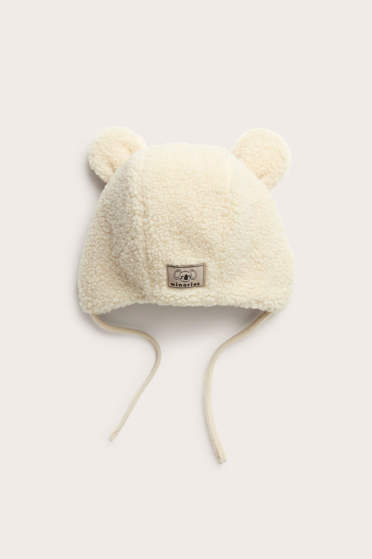 Framifrån, enfärgad beige pile babymössa med öron och knytsnören.