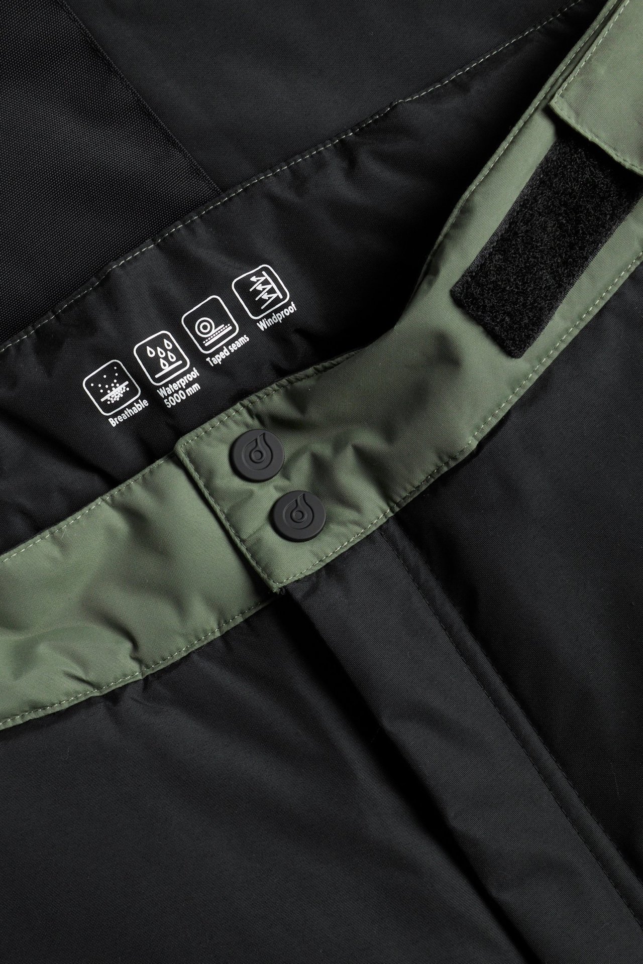 Zbliżenie na zielono-czarne spodnie snowboardowe w bloki kolorów z czarnymi guzikami i rzepem w pasie.