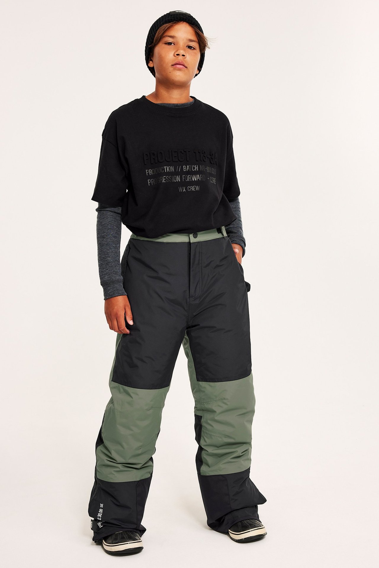 Widok z przodu dziecka w luźnych, blokowo wzorzystych czarno-zielonych spodniach snowboardowych, czarnym t-shircie i czapce.