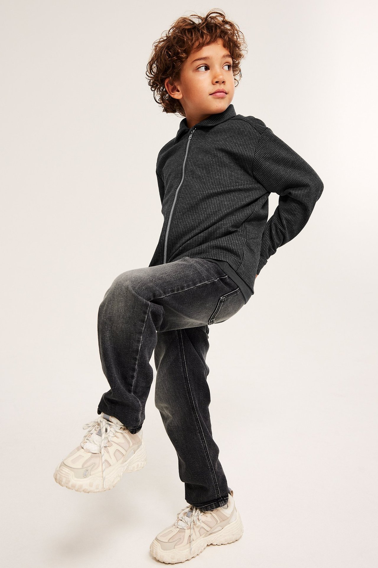Frontvy av ett barn i mörkgrå hundtandsmönstrad overshirt med dragkedja, mörkgrå jeans och ljusa sneakers.