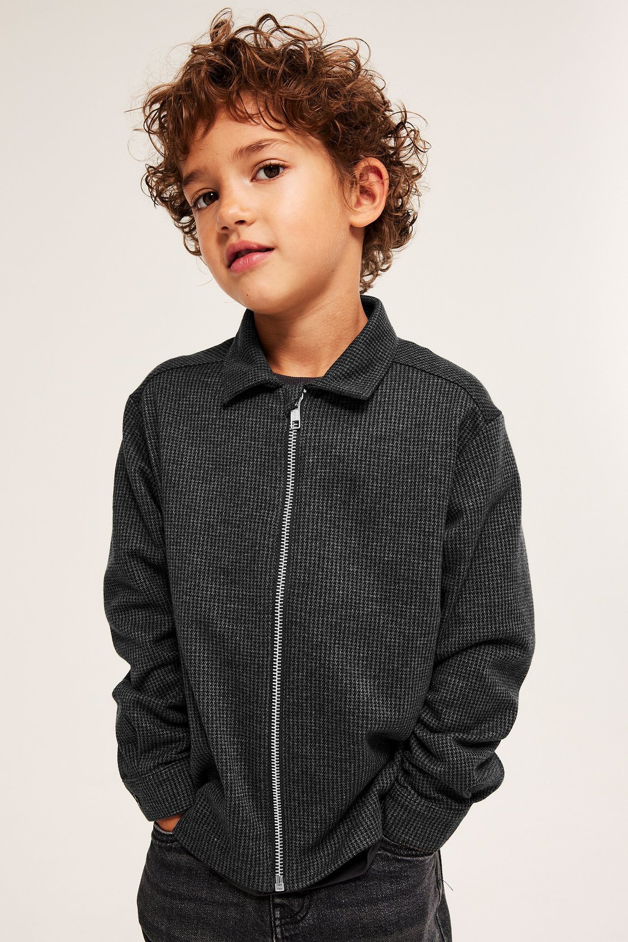 Frontbild av ett barn i mörkgrå hundtandsmönstrad overshirt med dragkedja.