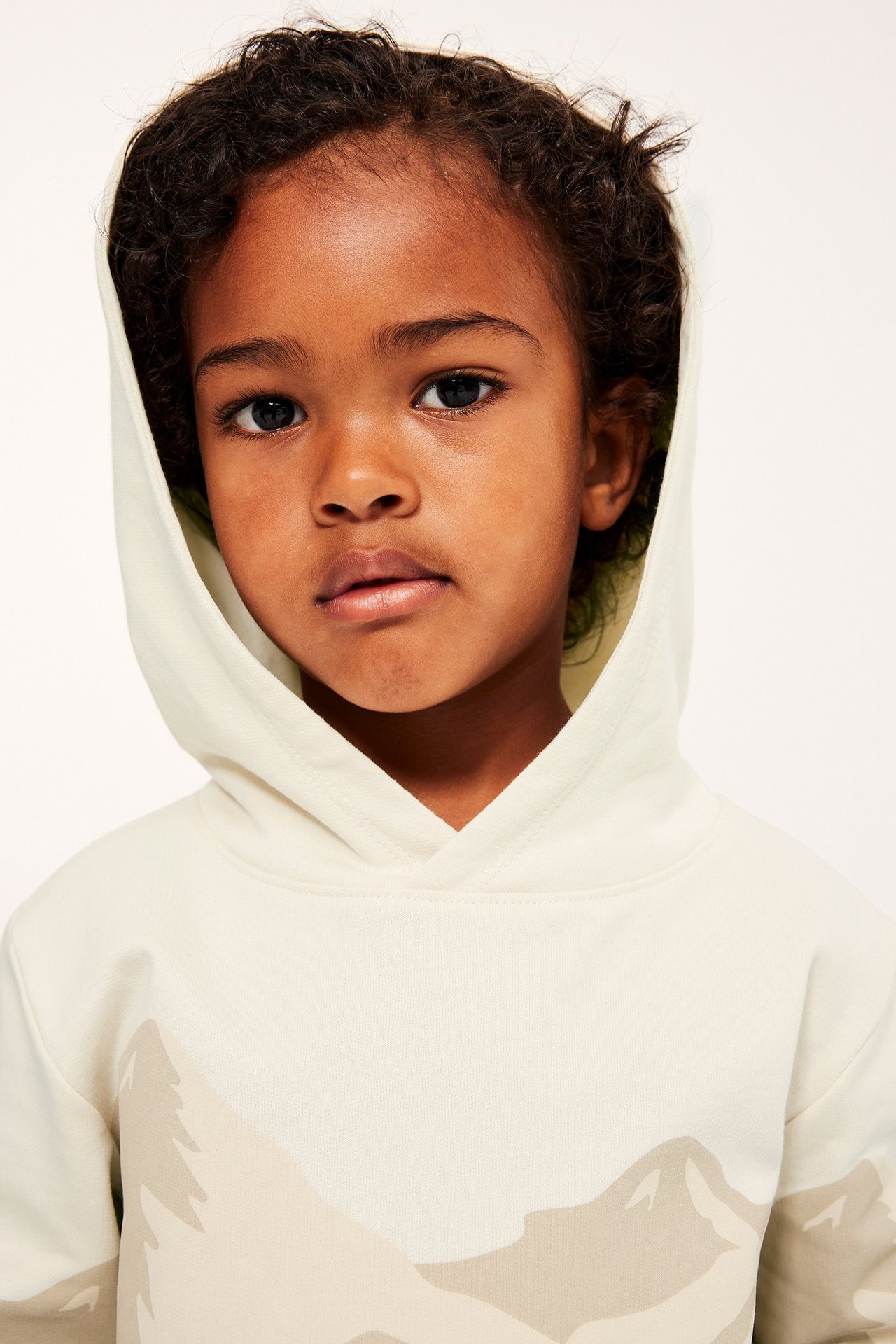 Nærbilde forfra av et barn i en lys beige bomullshettgenser med diskret fjell- og skogmønster.