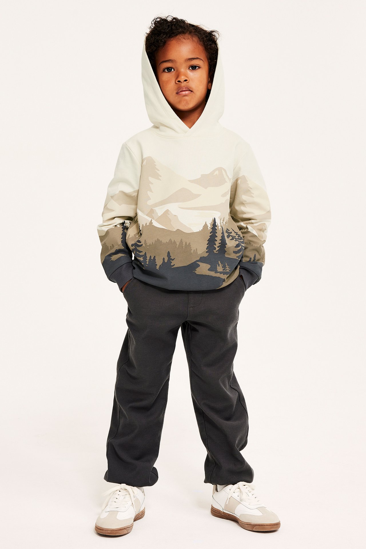Framifrån, ett barn i en ljusbeige mönstrad hoodie med berg, mörkgrå byxor och vita sneakers.