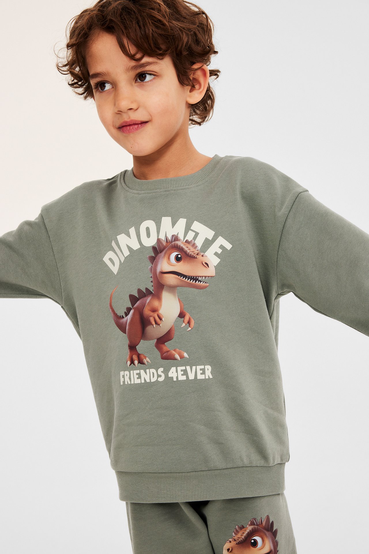 Frontvy av ett barn i en grön bomullssweatshirt med dinosaurietryck och matchande gröna sweatpants.