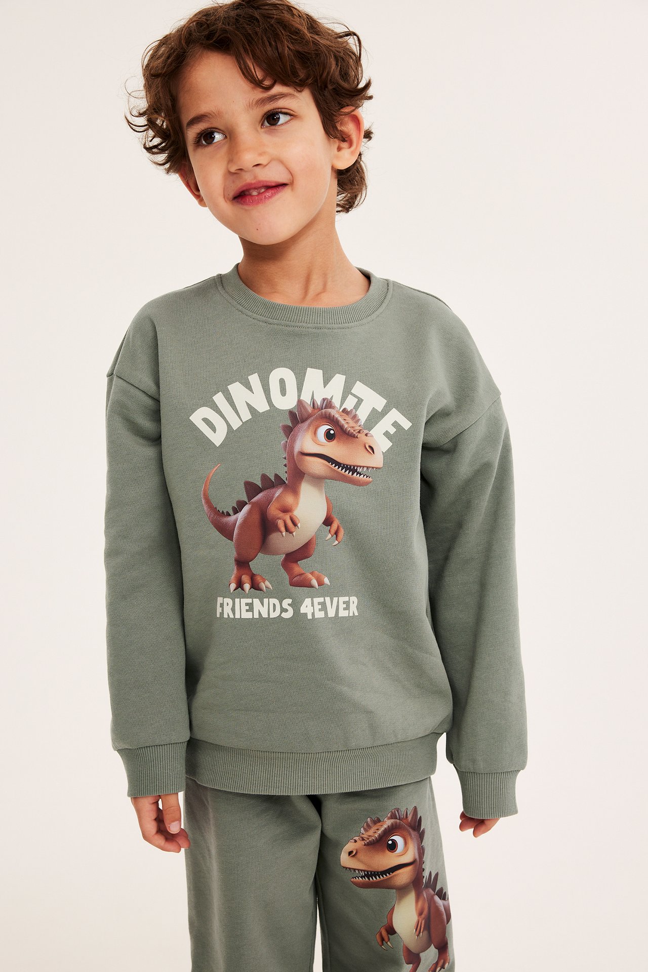 Framifrån: Ett barn i grön bomullssweatshirt med dinosaurietryck, matchande gröna sweatpants.