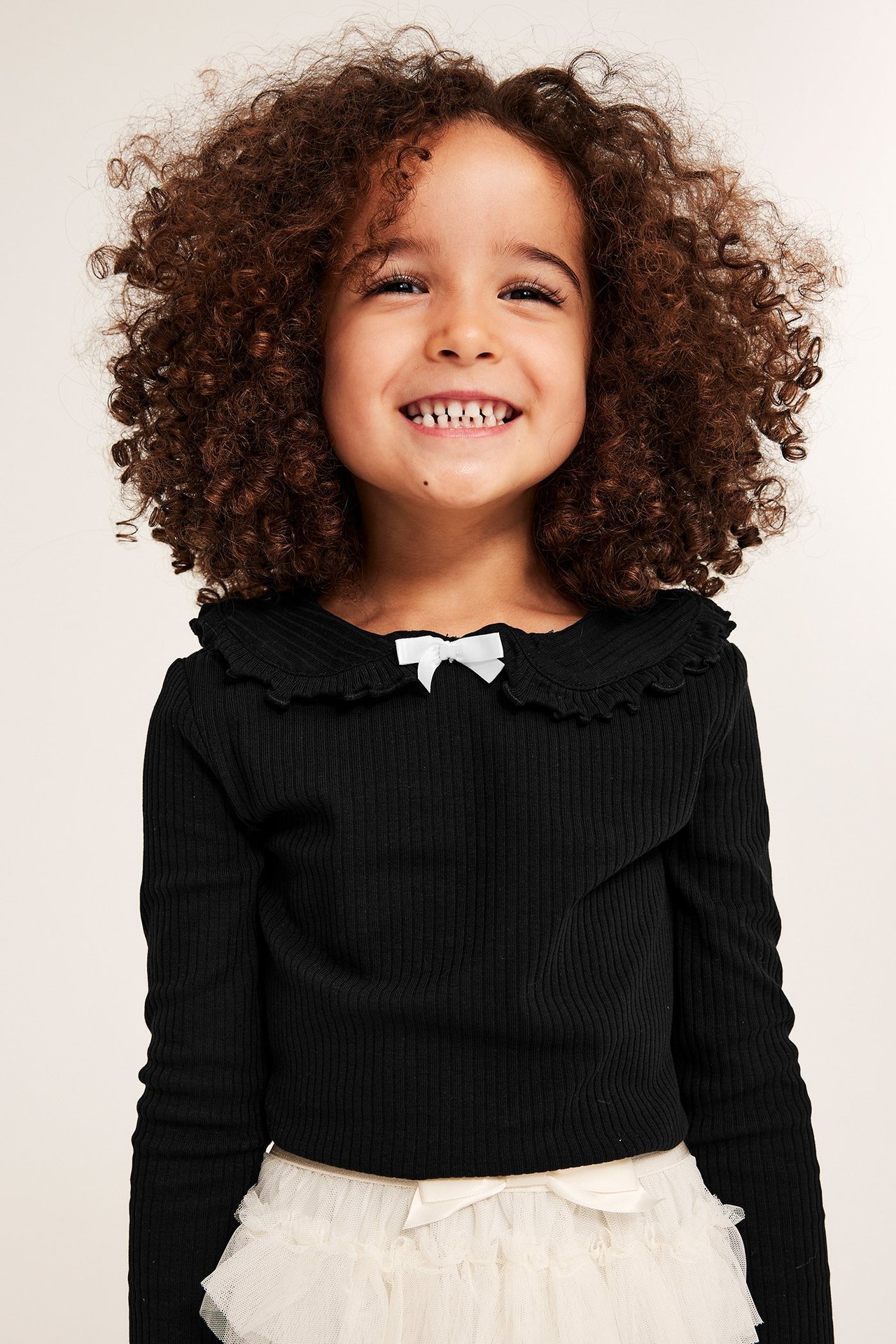 Leende barn i svart ribbad topp med volangkrage, vit rosett, och en krämfärgad tyllkjol. Framifrån.