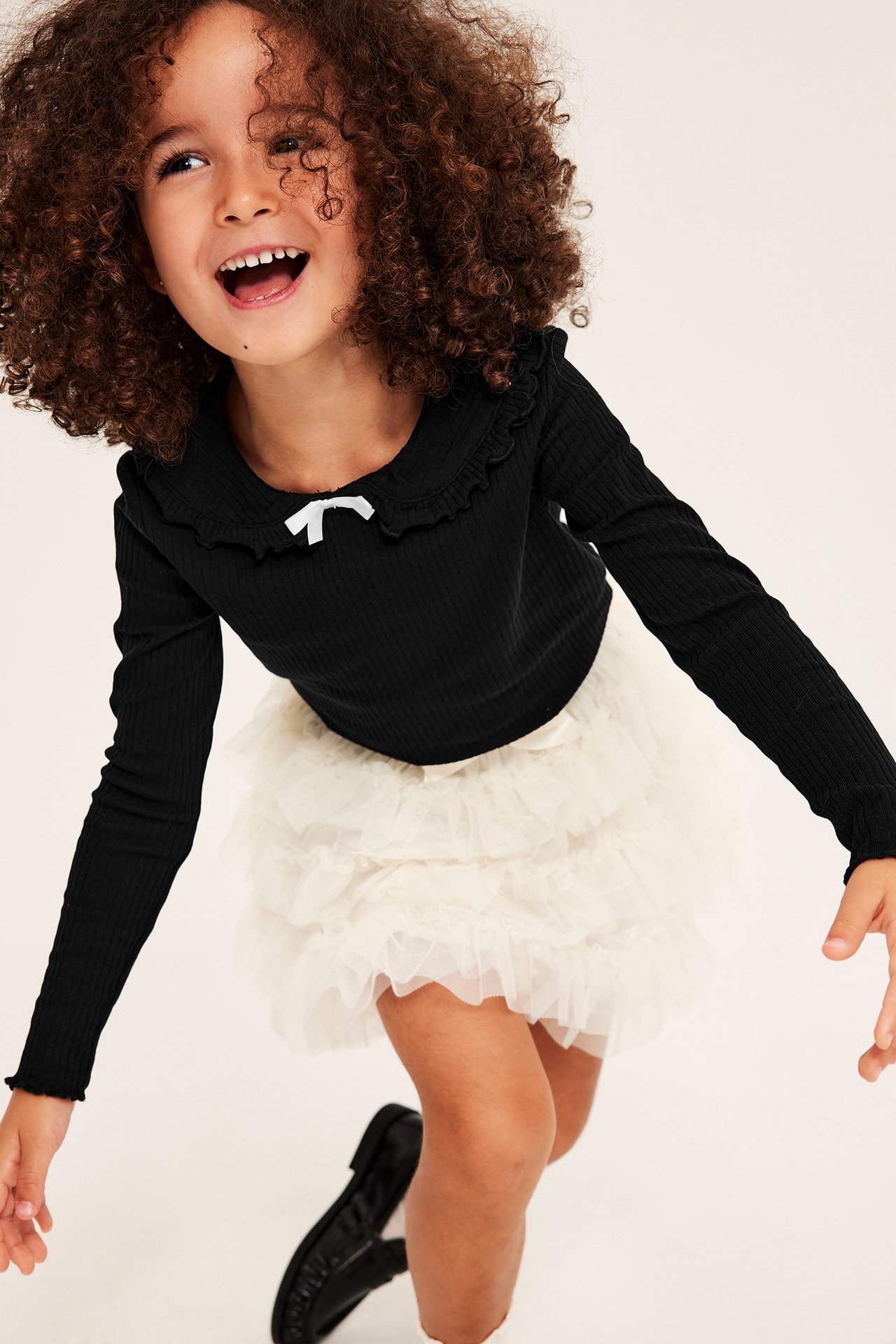 Framifrån: Barn bär svart ribbad topp med volangkrage, vit rosett och gräddfärgad tyllkjol.