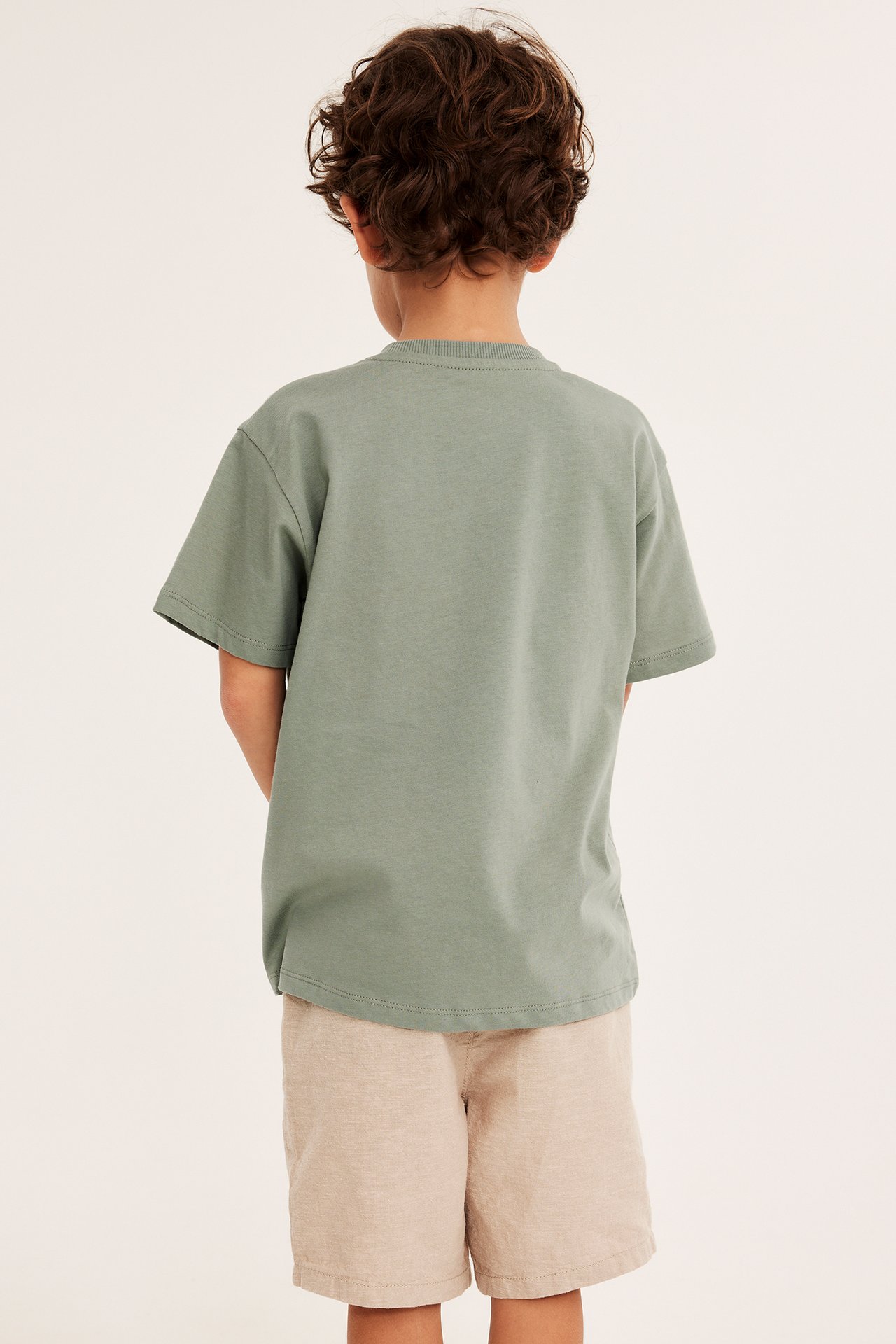Bakfra, et barn i en mørkegrønn bomulls-t-skjorte med ribbet hals og lysebeige shorts.