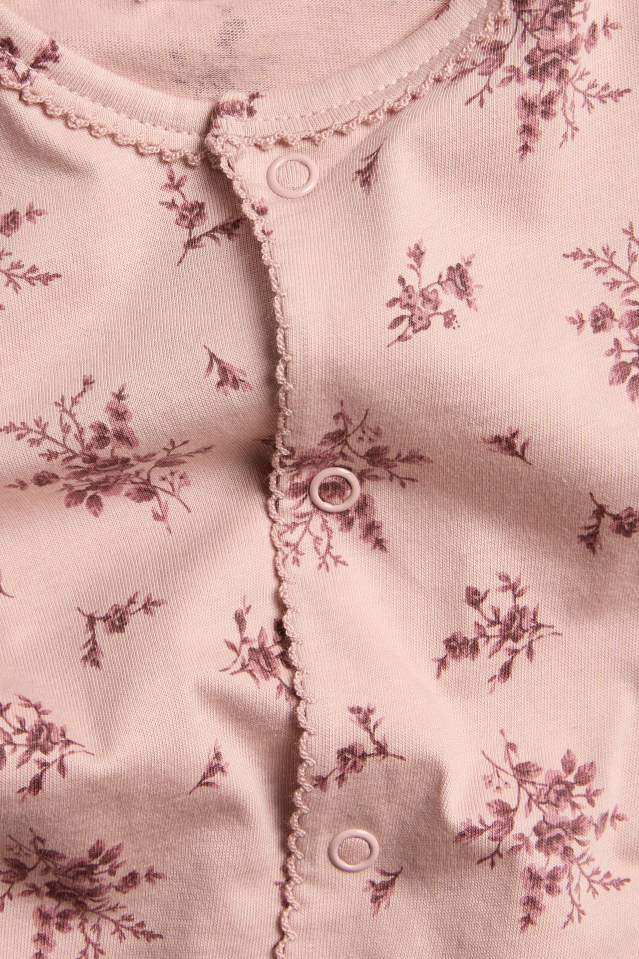 Lähikuva edestä: vaaleanpunainen puuvillainen vauvanpyjama, jossa kukkakuvio, nepparit ja picot-reuna.
