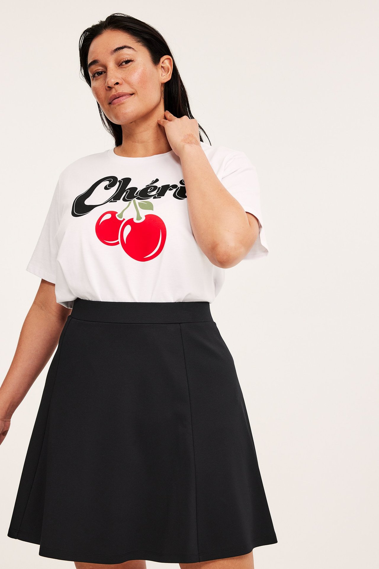 Widok z przodu: kobieta w czarnej rozkloszowanej spódnicy skater i białym t-shircie z nadrukiem "Chérie" wiśnie.