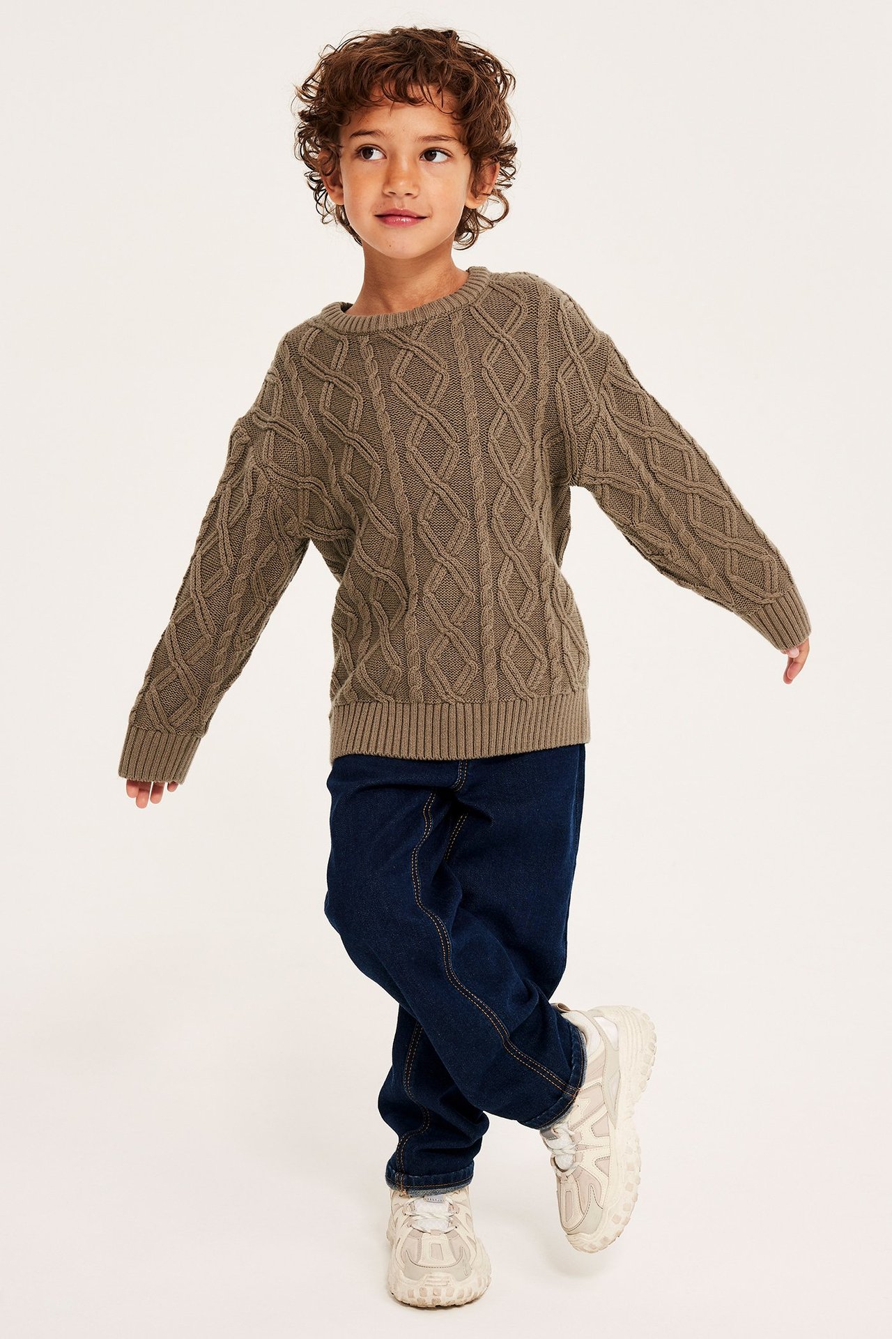 Edestäpäin, lapsi yllään ruskea palmikkoneulepusero, tummansiniset farkut ja vaaleat paksupohjaiset lenkkarit.