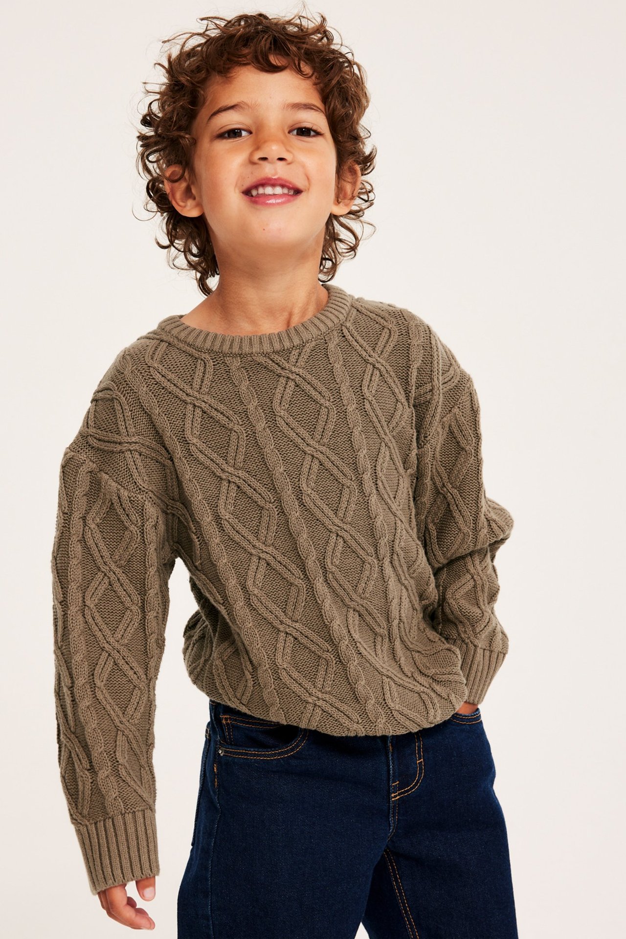 Widok z przodu uśmiechniętego dziecka w brązowym swetrze w warkocze i ciemnoniebieskich dżinsach.