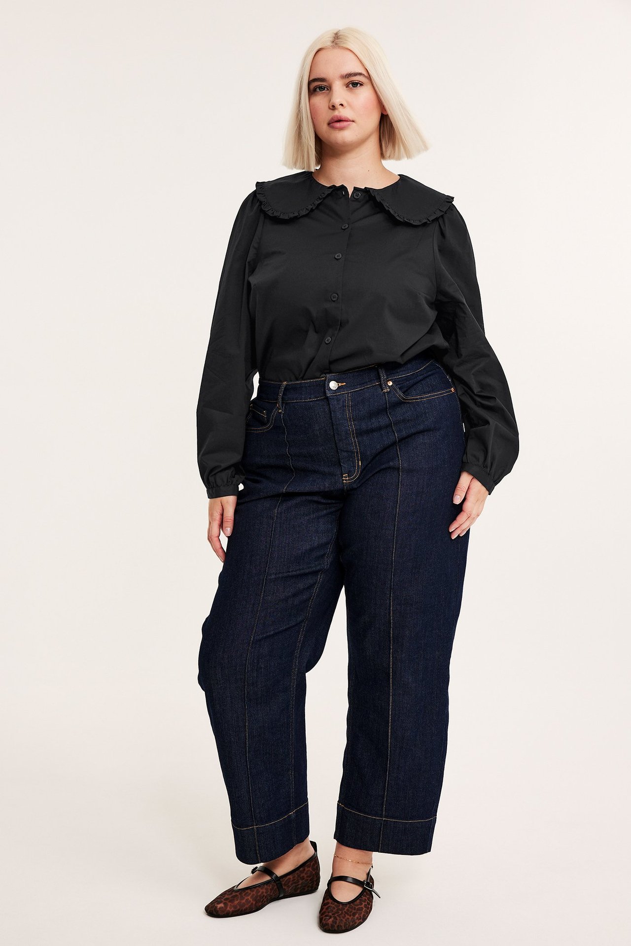 Framifrån: kvinna i svart bomullsblus med rundad krage, mörkblå vida jeans, leopardmönstrade ballerinaskor.