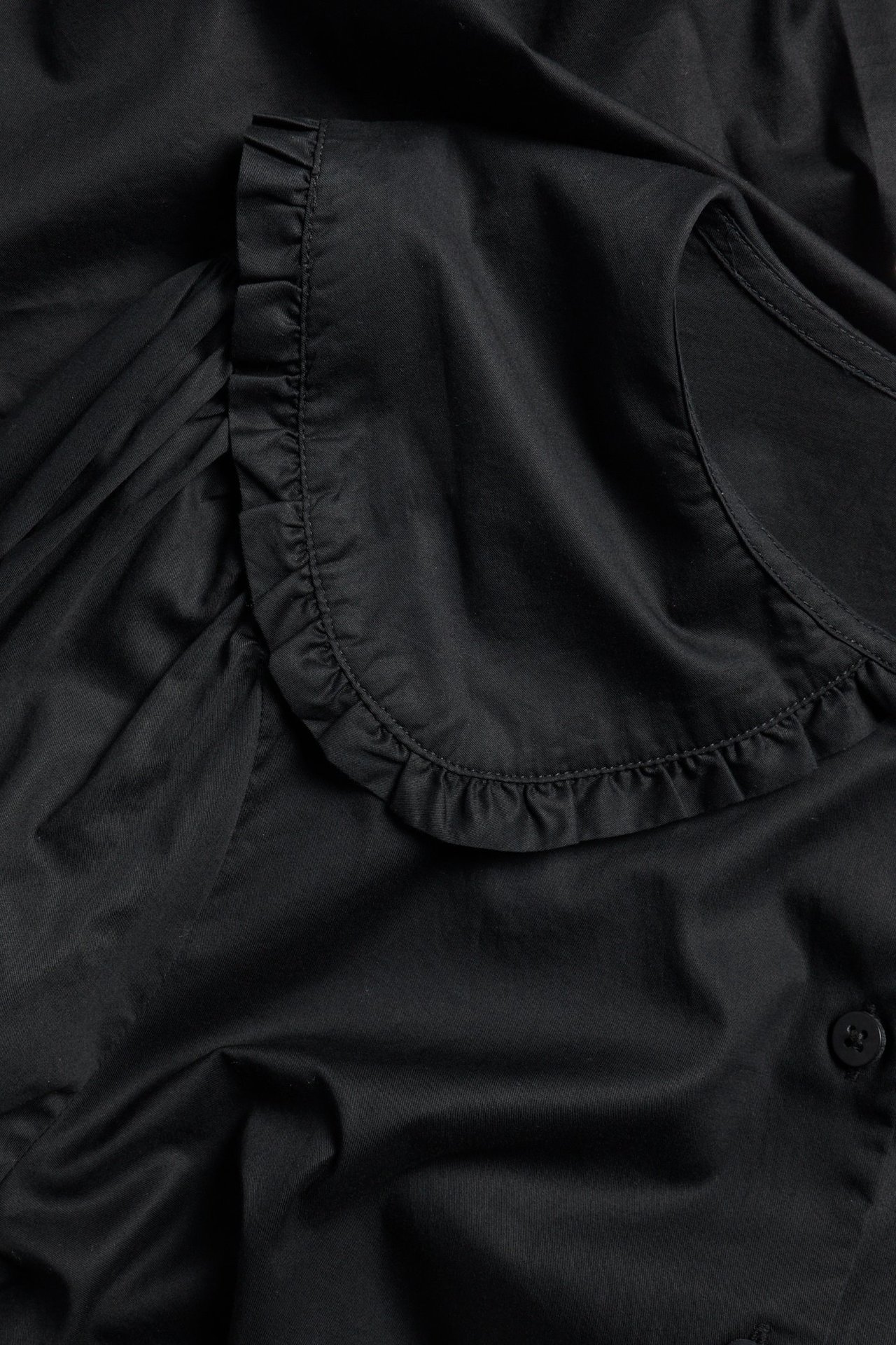 Zbliżenie czarnej bawełnianej bluzki z falbankowym, zaokrąglonym kołnierzem i widocznym guzikiem.