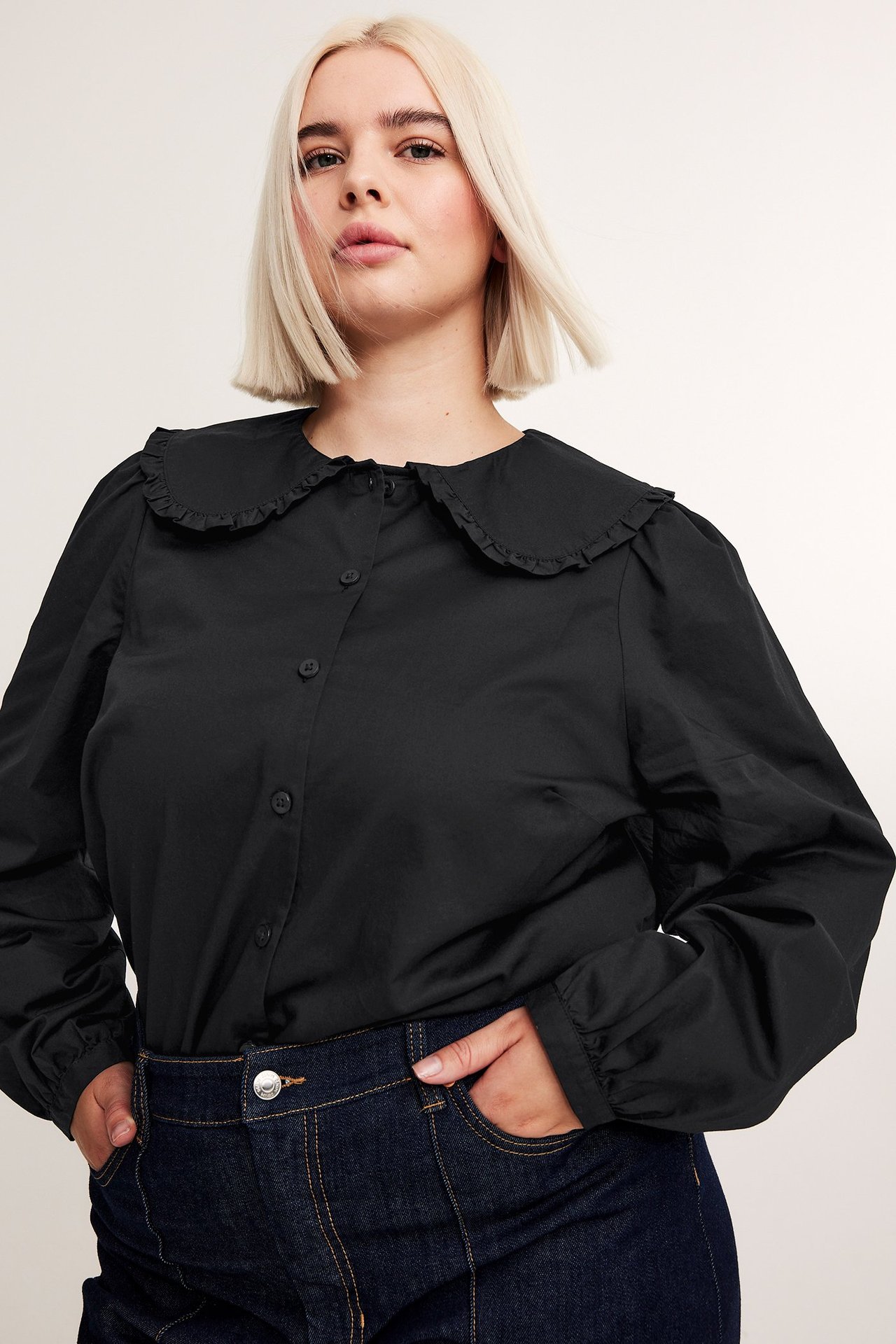 Framifrån: kvinna i svart bomullsblus med volangkrage och långa ärmar, samt mörkblå jeans.
