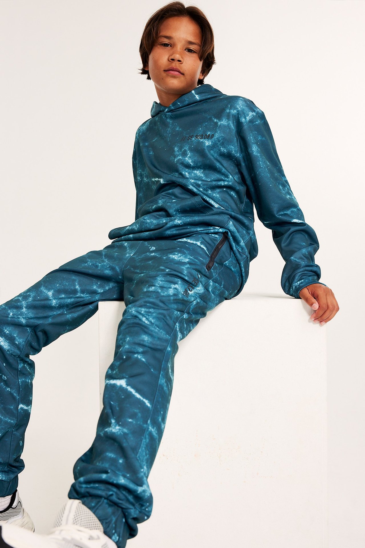 Z przodu: chłopiec w zielonych/turkusowych wzorzystych spodniach dresowych i pasującej bluzie.