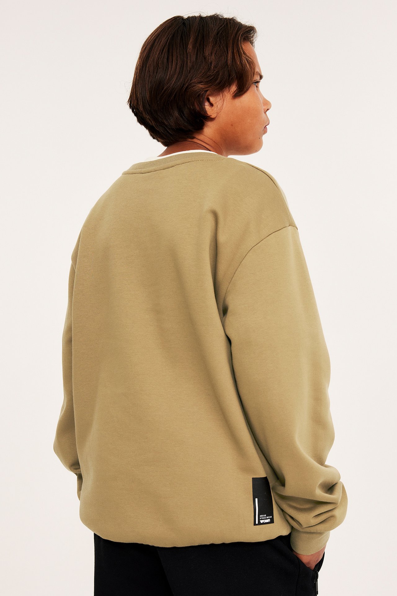 Taka-/sivukuva lapsesta yllään ruskea oversized collegepaita mustalla etiketillä ja mustat housut.