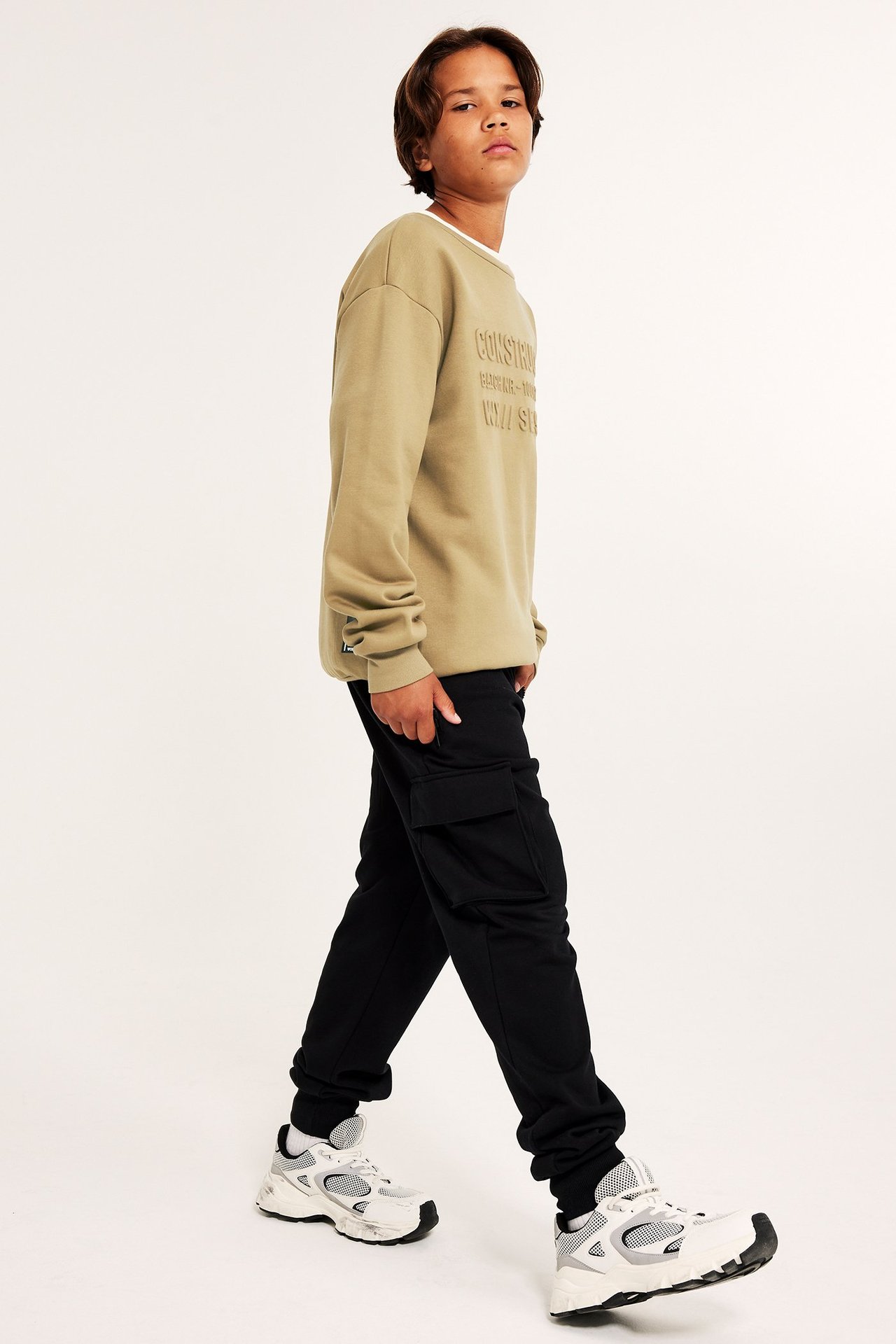 Widok z boku chłopca w brązowej bluzie z nadrukiem 3D, czarnych spodniach cargo i białych sneakersach.