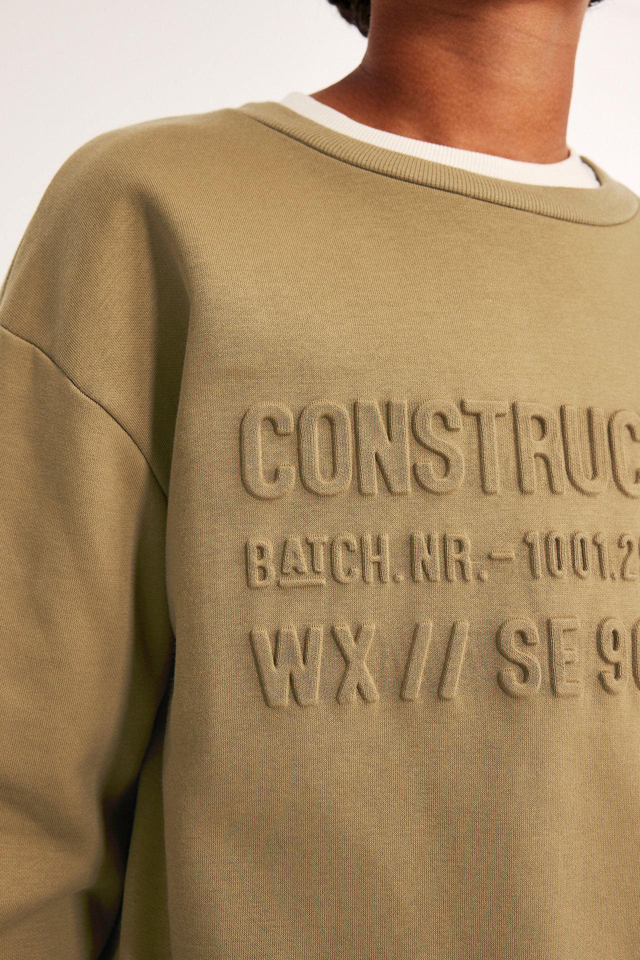 Lähikuva edestä: ruskea puuvillasekoitesweatshirt kohokuvioidulla "CONSTRUC"-tekstillä, valkoisen topin päällä.