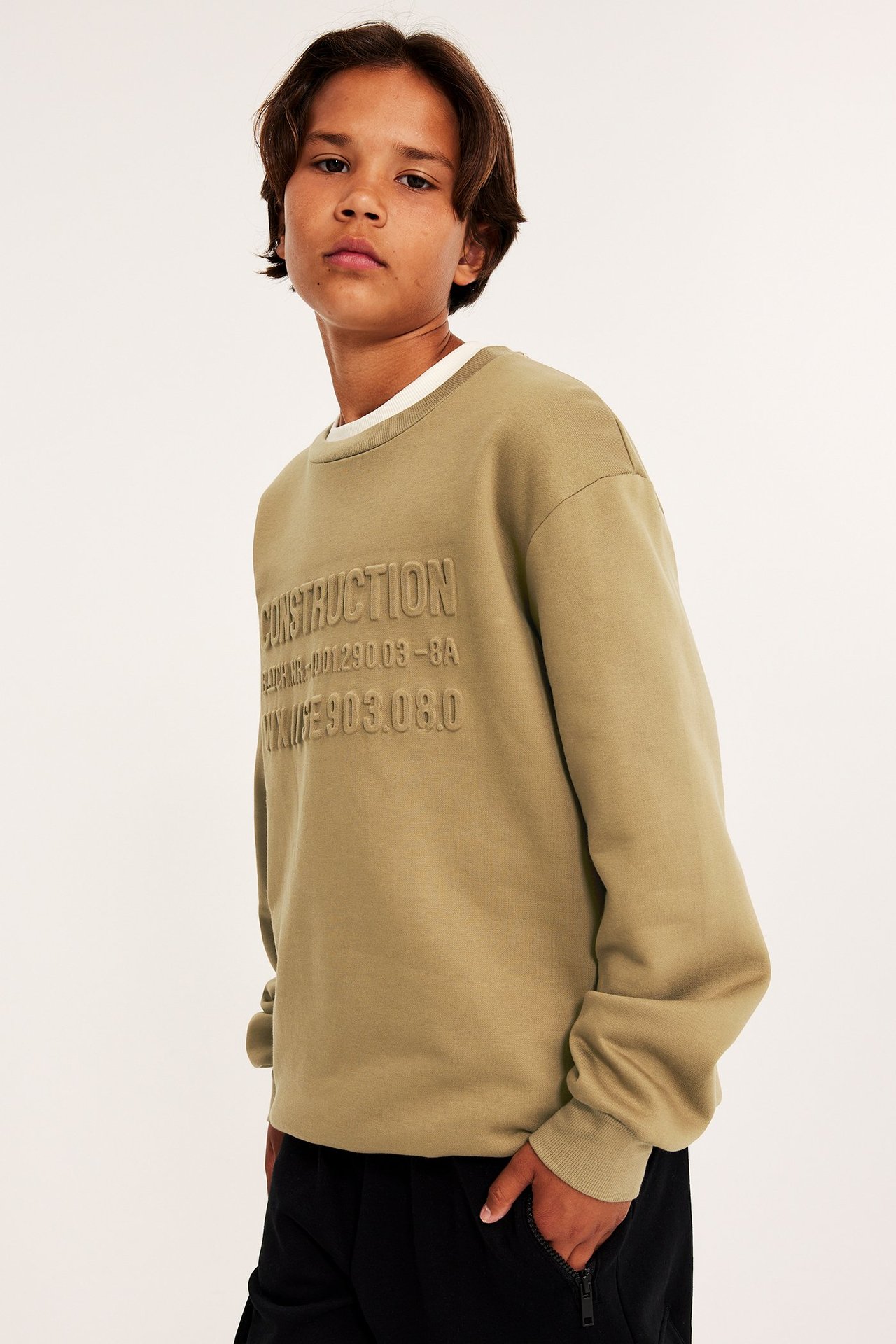Dziecko z przodu: brązowa bluza z wytłoczonym tekstem, biały t-shirt i czarne spodnie.