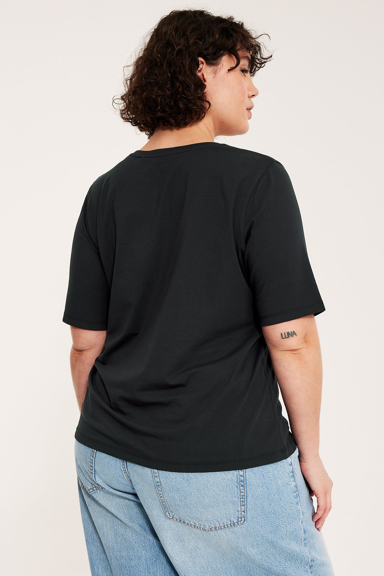Widok od tyłu kobiety w czarnym, jednolitym bawełnianym T-shircie i jasnoniebieskich luźnych dżinsach.