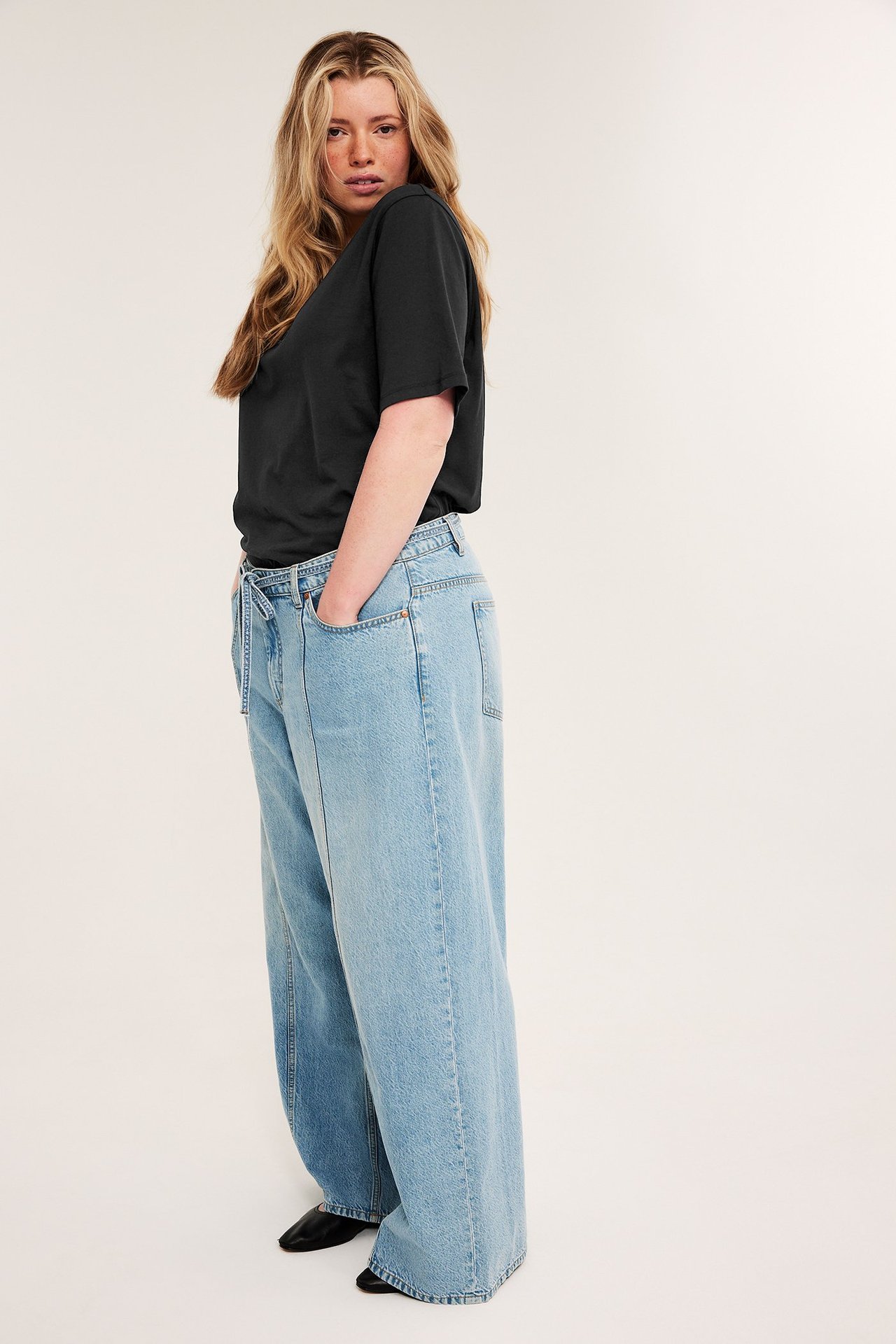 Sidevisning av en kvinne i svart figurnær bomulls-t-skjorte, lyseblå vide jeans og svarte flate sko.
