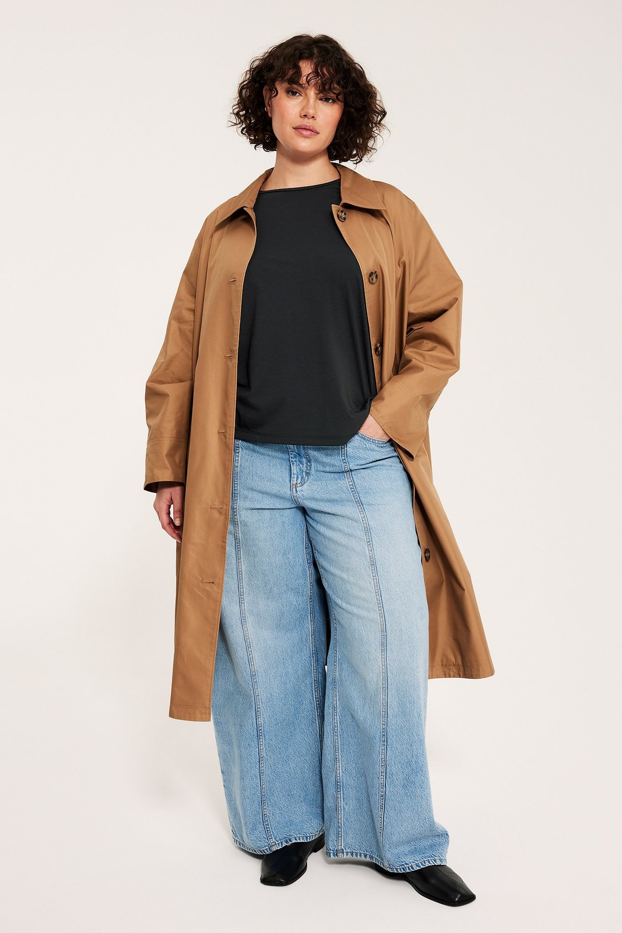 Kvinna bär svart T-shirt, lång brun trenchcoat, ljusblå vida jeans och svarta skor.