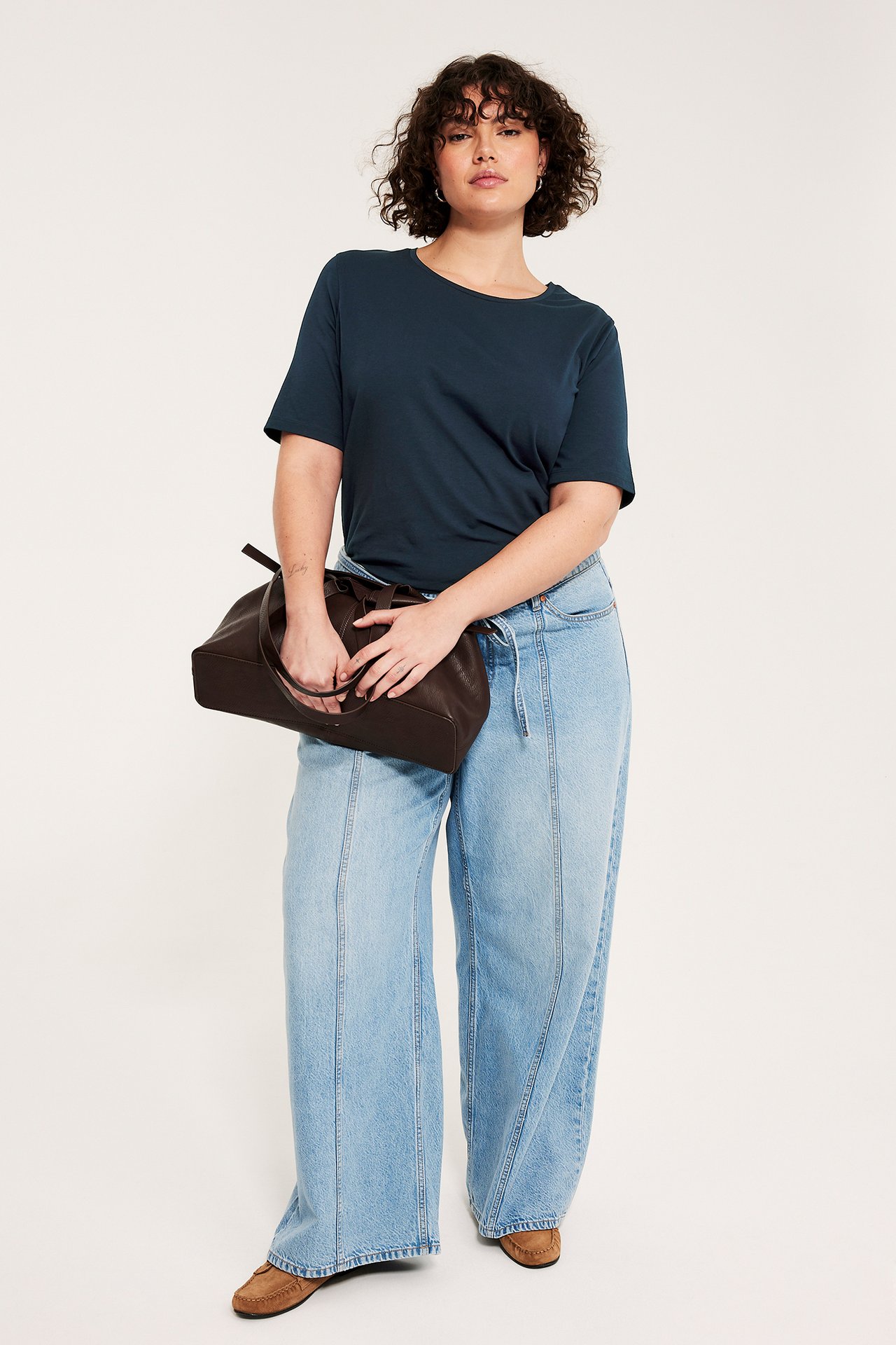 Frontal: Kvinne i mørk blå t-skjorte, lyseblå vide jeans, holder en brun håndveske.