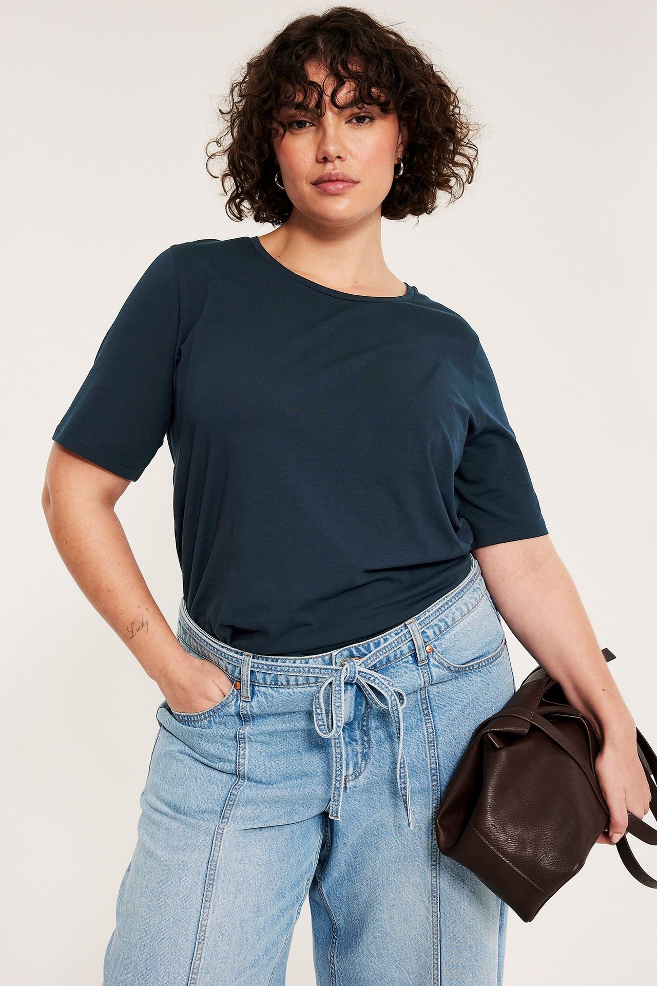 Frontutsikt av kvinne i mørkeblå t-skjorte, lyseblå jeans med knytebånd i midjen, holder en mørkebrun veske.