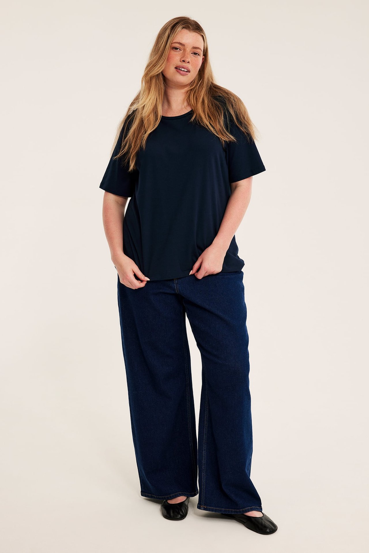 Frontbilde av kvinne i marineblå t-skjorte, mørkeblå vide jeans og svarte ballerinasko.