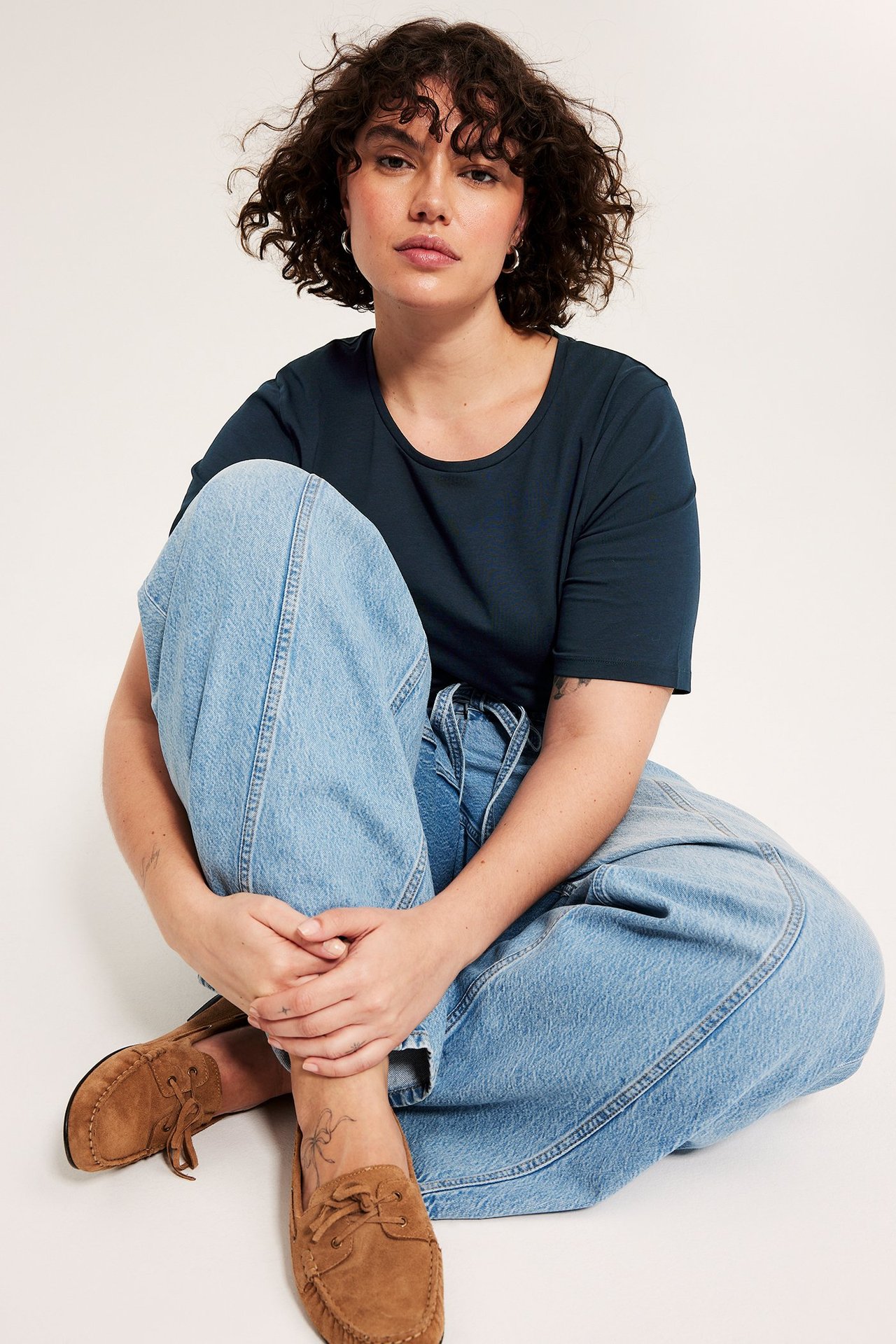 Z przodu: Kobieta w granatowej bawełnianej koszulce, jasnoniebieskich szerokich jeansach i brązowych mokasynach.