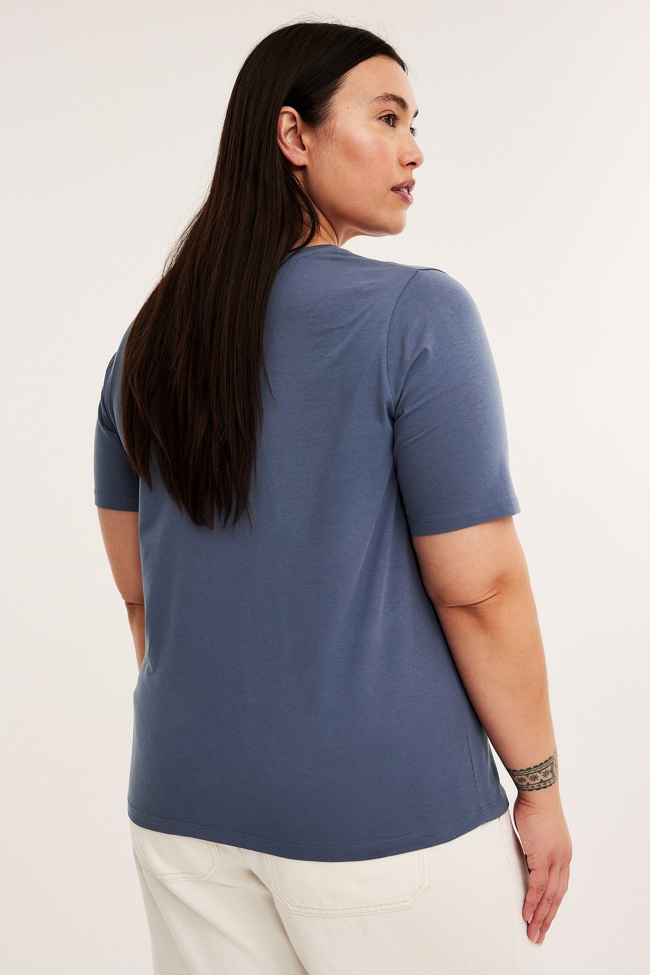Bak-/sidevisning av kvinne i blå kortermet t-skjorte i bomullsblanding og lyse bukser.