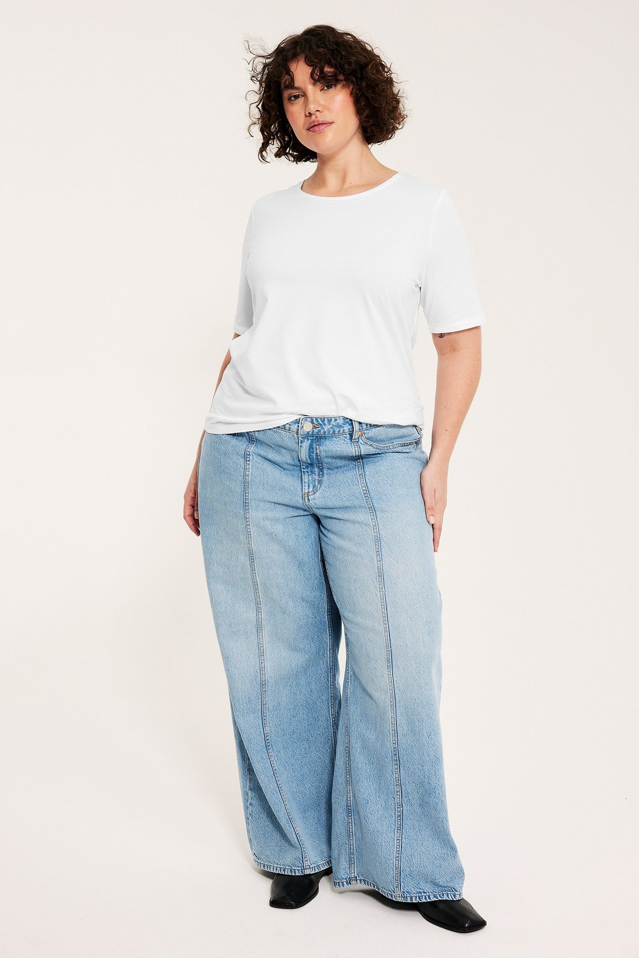 Edestä näkymä: nainen yllään valkoinen lyhythihainen T-paita ja vaaleansiniset leveälahkeiset farkut.