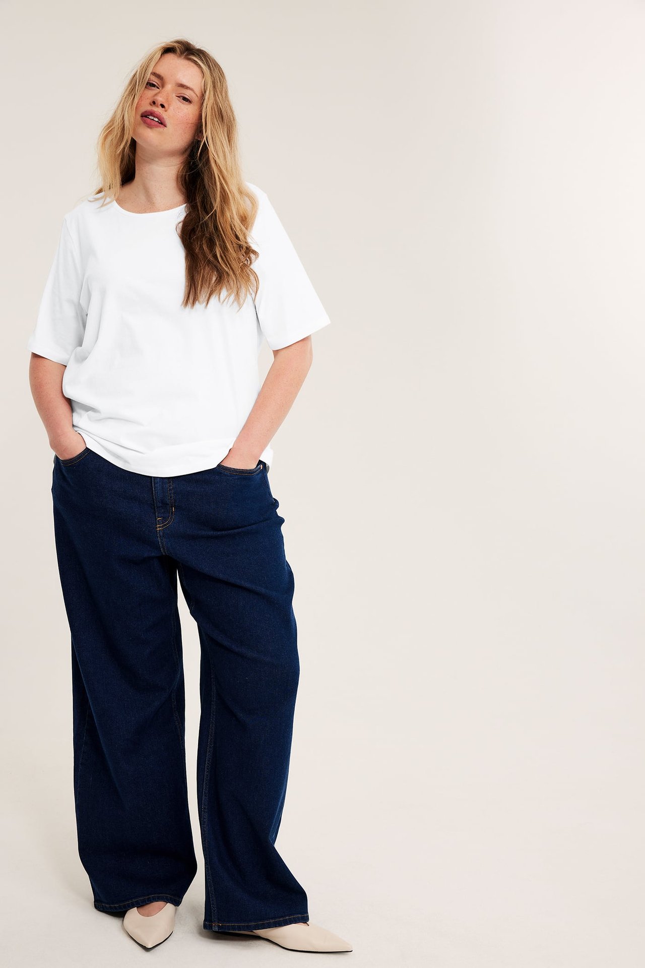 Kvinne i hvit ettersittende t-skjorte, mørkeblå vide jeans og lysebeige spisse ballerinasko.