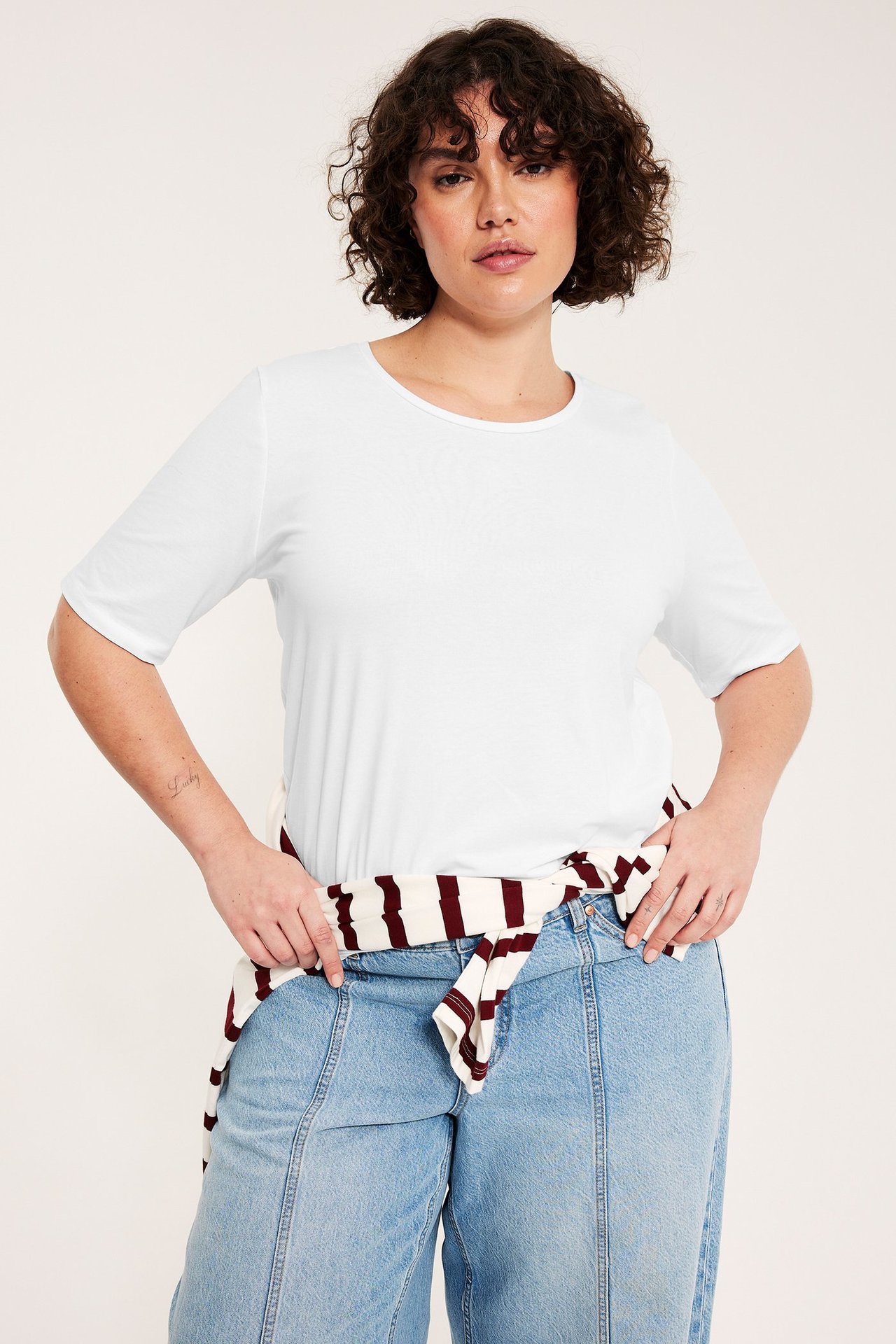 Kobieta w białym t-shircie, jasnoniebieskich jeansach i związanej w pasie bluzce w paski.