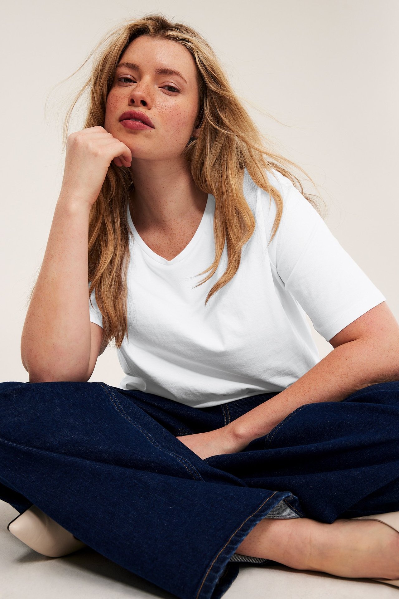 Forfra: Sitter kvinne i en hvit V-hals t-skjorte og mørkeblå vide jeans.