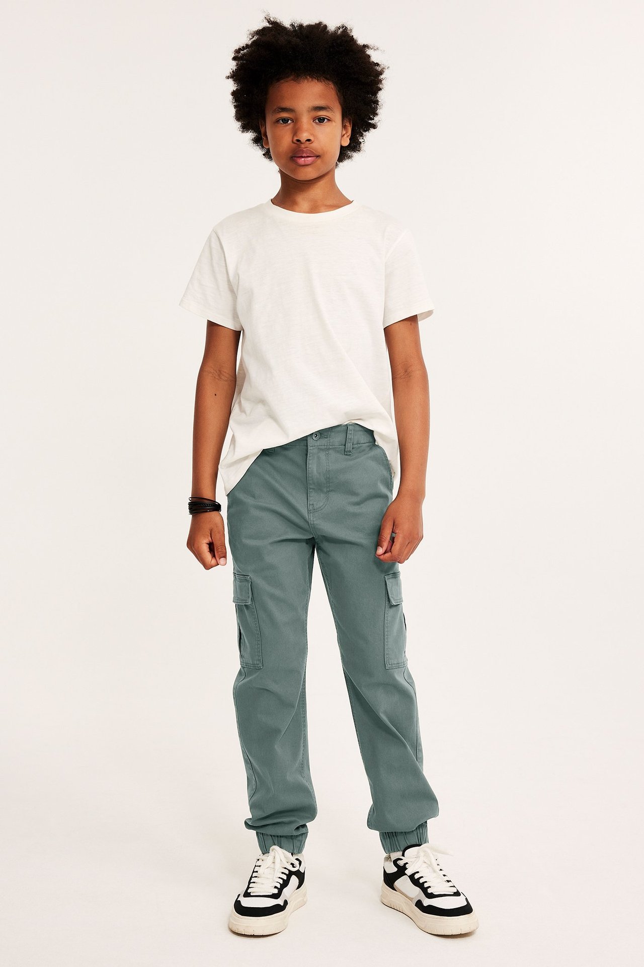 Widok z przodu: Dziecko w spodniach cargo khaki z kieszeniami, elastycznymi mankietami, białym t-shirtem i trampkami.