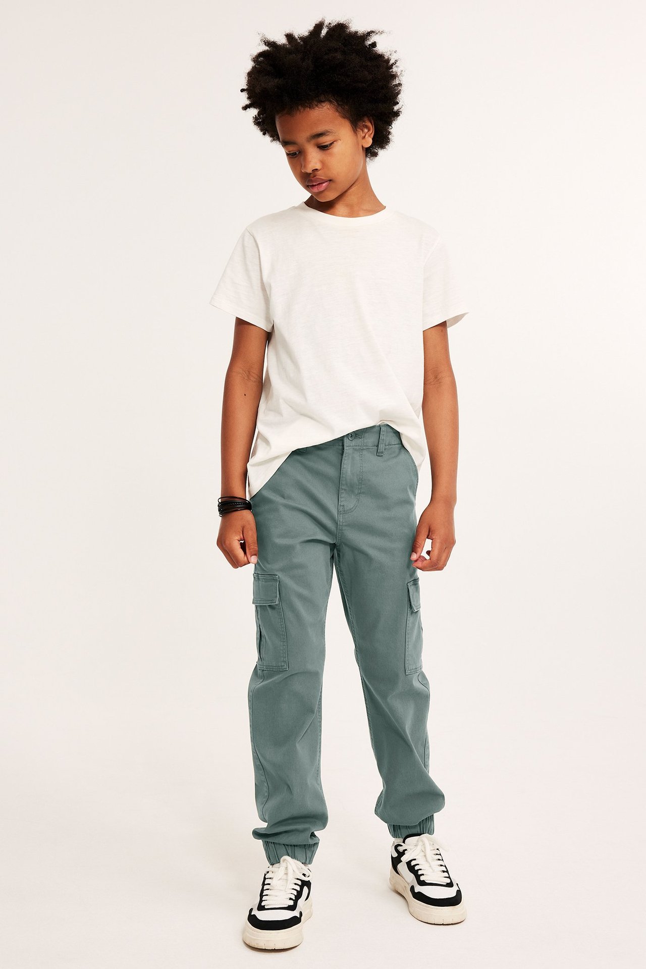 Widok z przodu: Dziecko w matowozielonych bawełnianych spodniach cargo z kieszeniami, białym t-shircie i sneakersach.