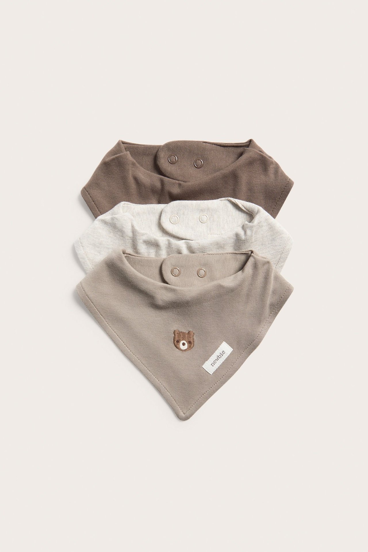 Edestä näkymä kolmesta pinotusta vauvan puuvillaruokalapusta: beige karhubrodeerauksella, vaaleanharmaa meleerattu ja ruskea, neppareilla.