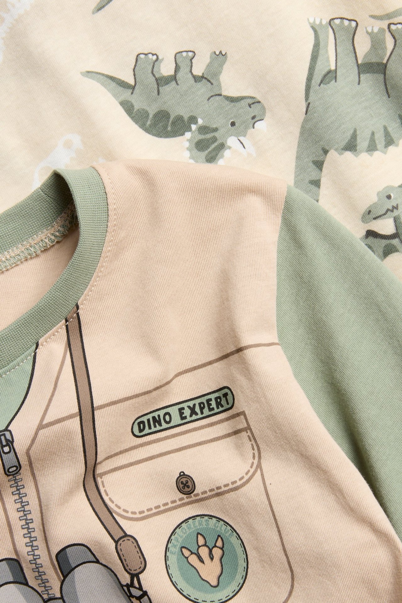 Zbliżenie na dwie dziecięce piżamy: jedna beżowa z nadrukiem 'Dino Expert', druga z zielonym wzorem dinozaurów.