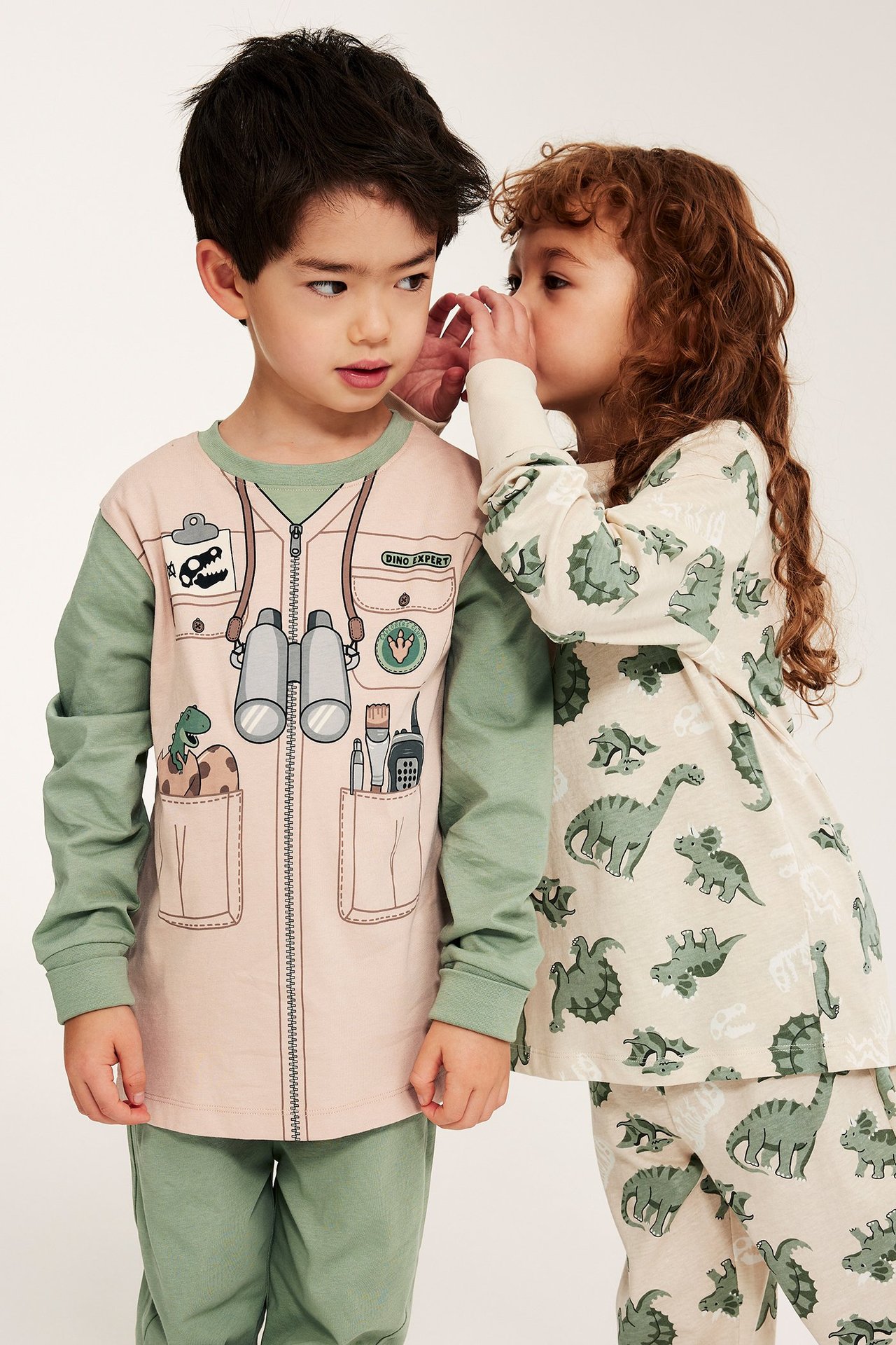 Edestä: lapsi vihreässä ja beigessä dinosaurus-tutkijapyjamassa, toinen lapsi beigessä dinosauruskuvioisessa setissä.