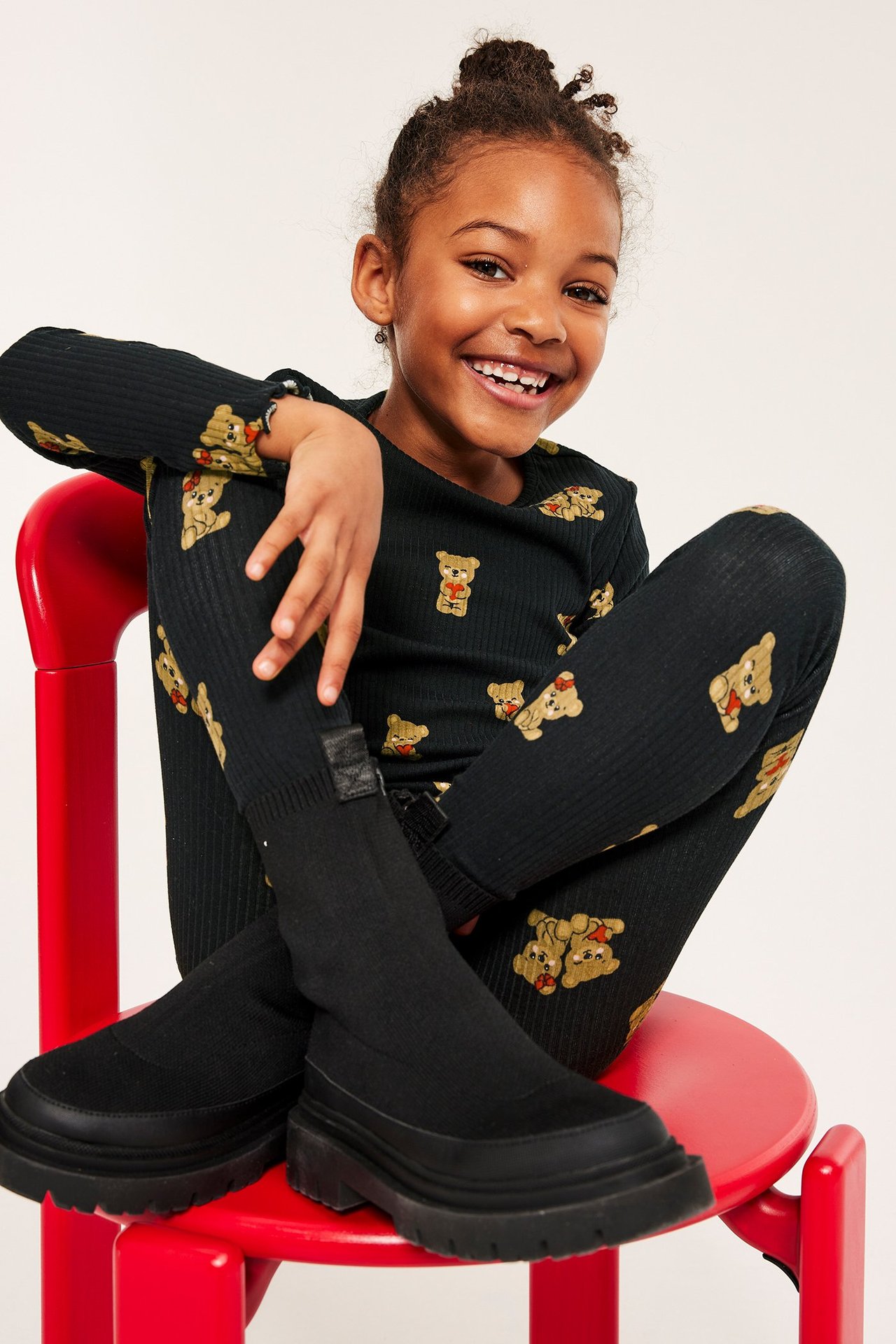 Dziewczynka w czarnej, wzorzystej bluzie z misiami, leggingsy i buty, siedzi, uśmiecha się.