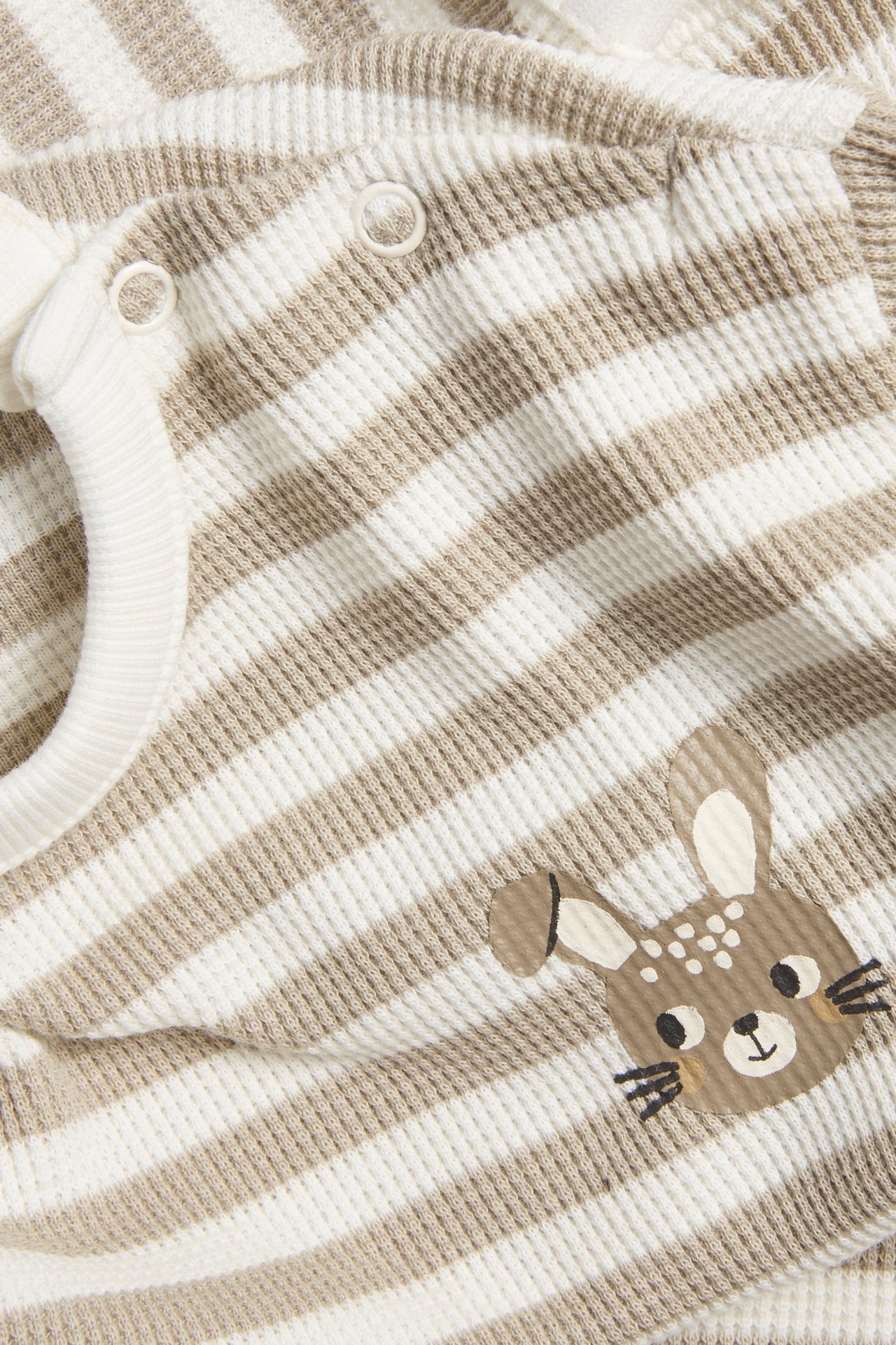 Zbliżenie na beżowo-białą, pasiastą bluzę niemowlęcą z dzianiny waflowej z zatrzaskami i aplikacją jelenia.