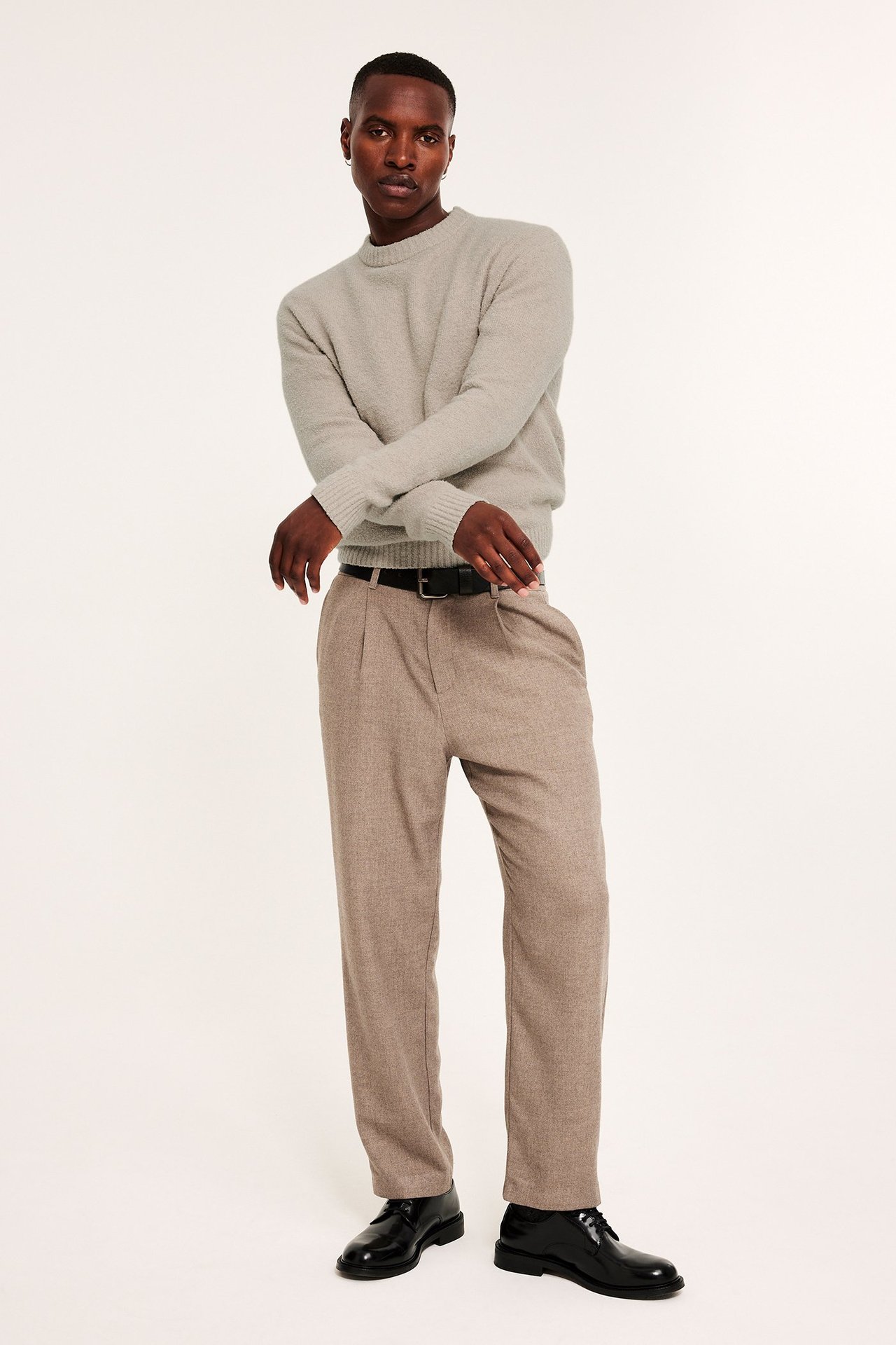 Frontbilde av mann i beige bouclé strikkegenser, brune plisserte bukser, svart belte og sko.