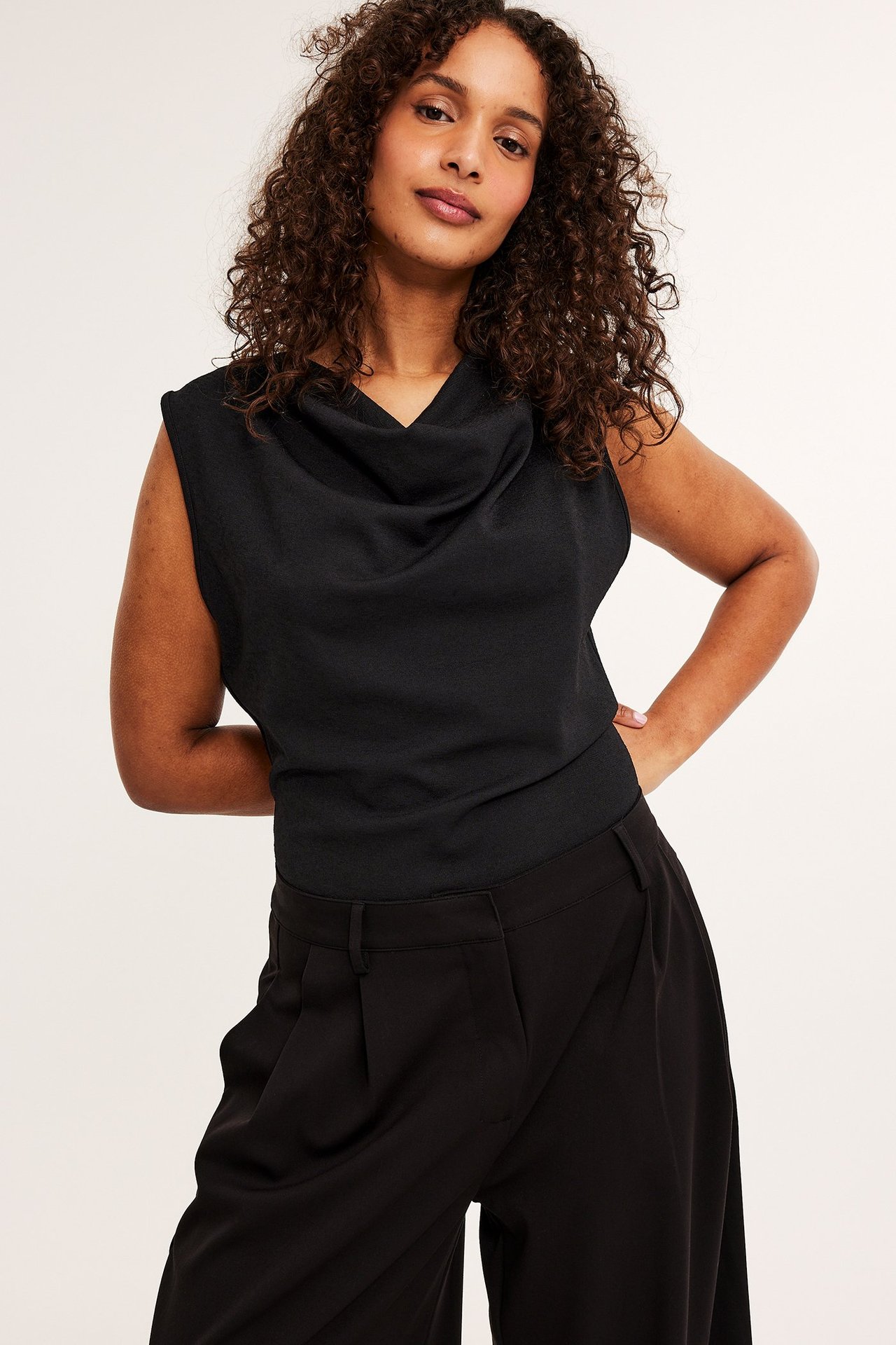 Forfra, kvinne i svart drapert ermeløs topp og svarte vide bukser.