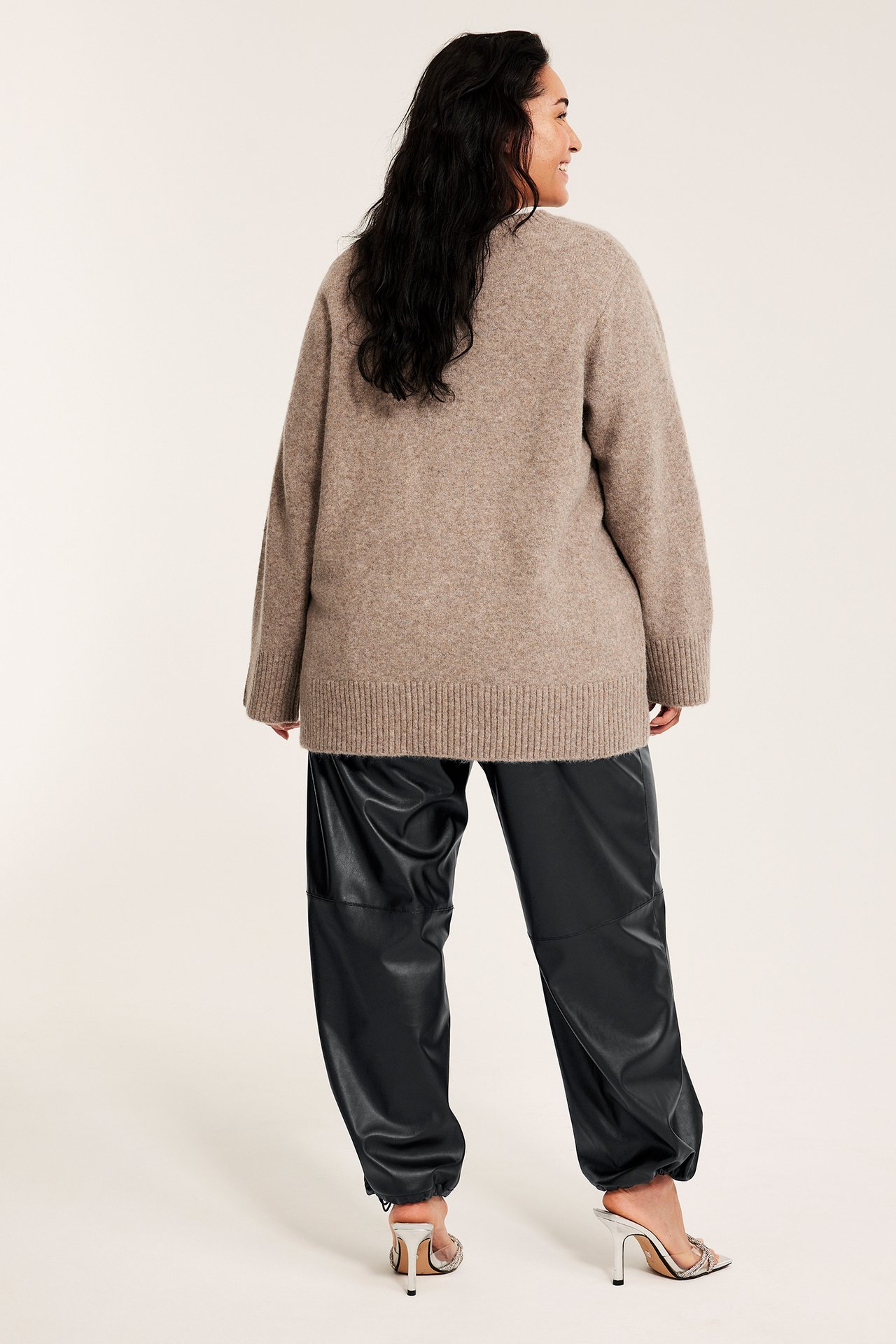 Bakifrån, kvinna i svarta PU pull-on byxor med resår i benslut, en beige stickad tröja och silvermules.