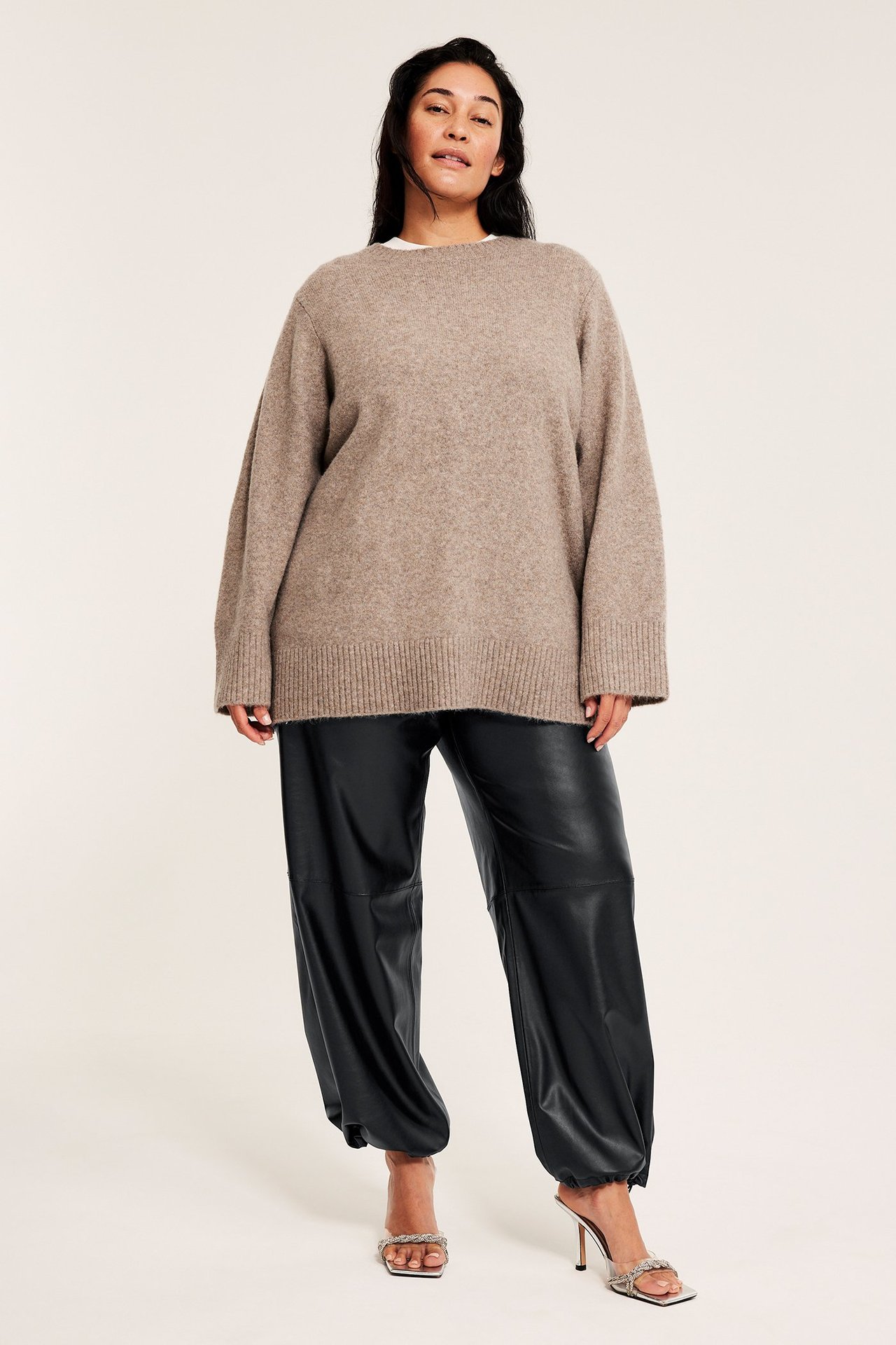 Edestäpäin nainen yllään mustat tekonahkaiset pull-on-housut, beige ylisuuri neule ja hopeiset korkokengät.