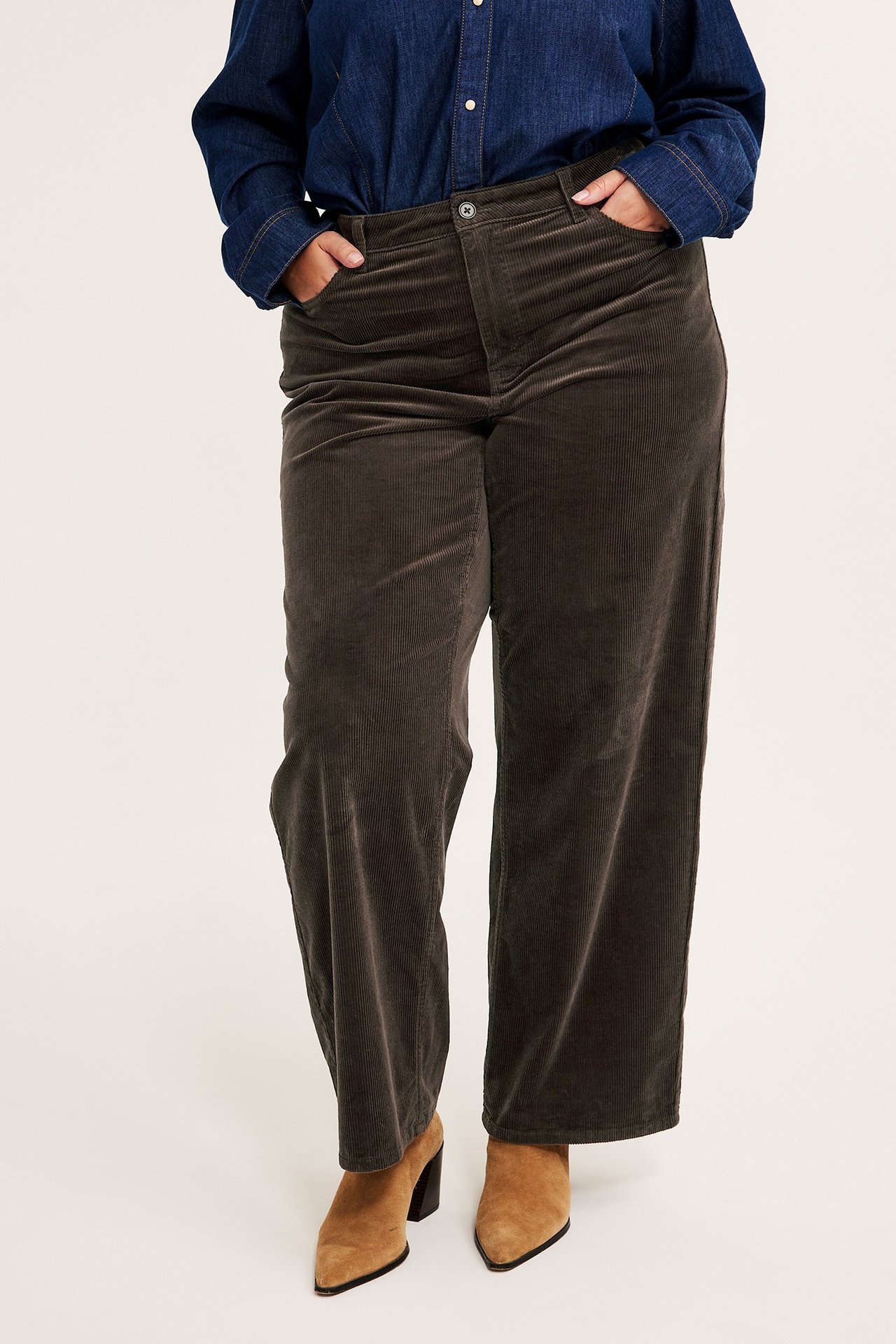 Framifrån: Kvinna i mörkbruna vida manchesterbyxor, en mörkblå jeansskjorta och ljusbruna klackstövlar.