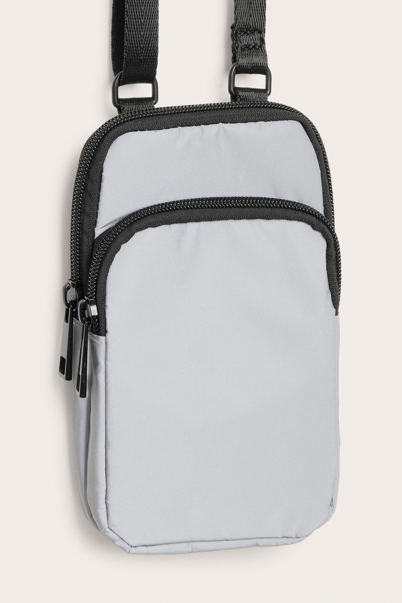 Frontvy av en silverfärgad reflekterande mobilväska med två dragkedjeförsedda fack och svart justerbar rem.